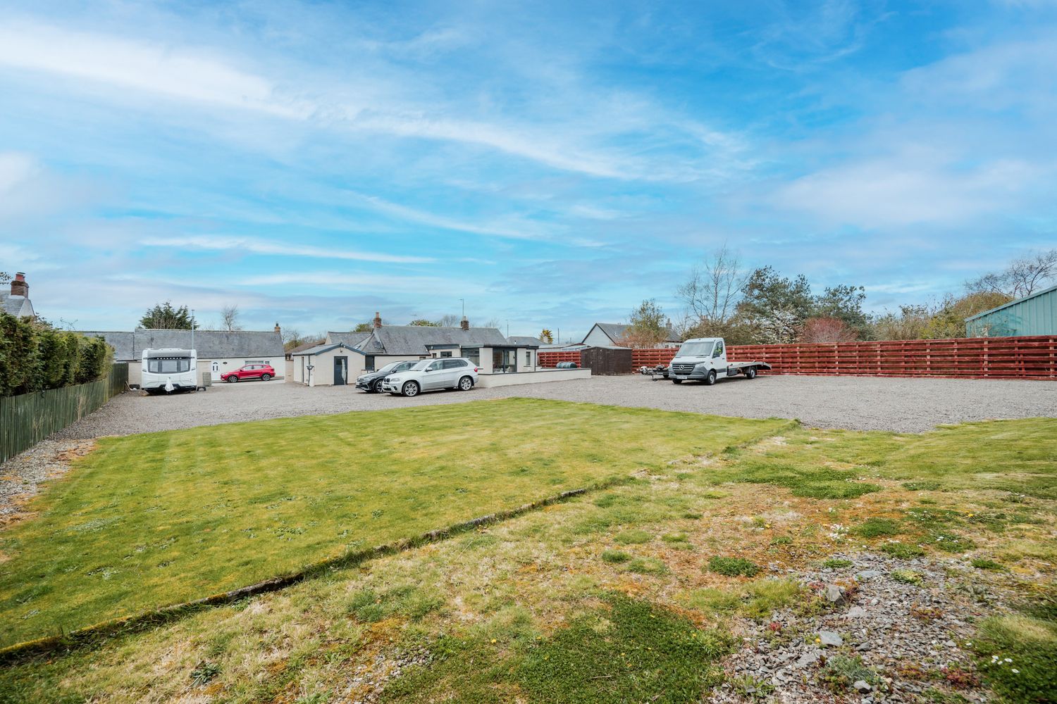 Large empty grass field with parked cars and buildings visible in the background under blue skies with wispy clouds.