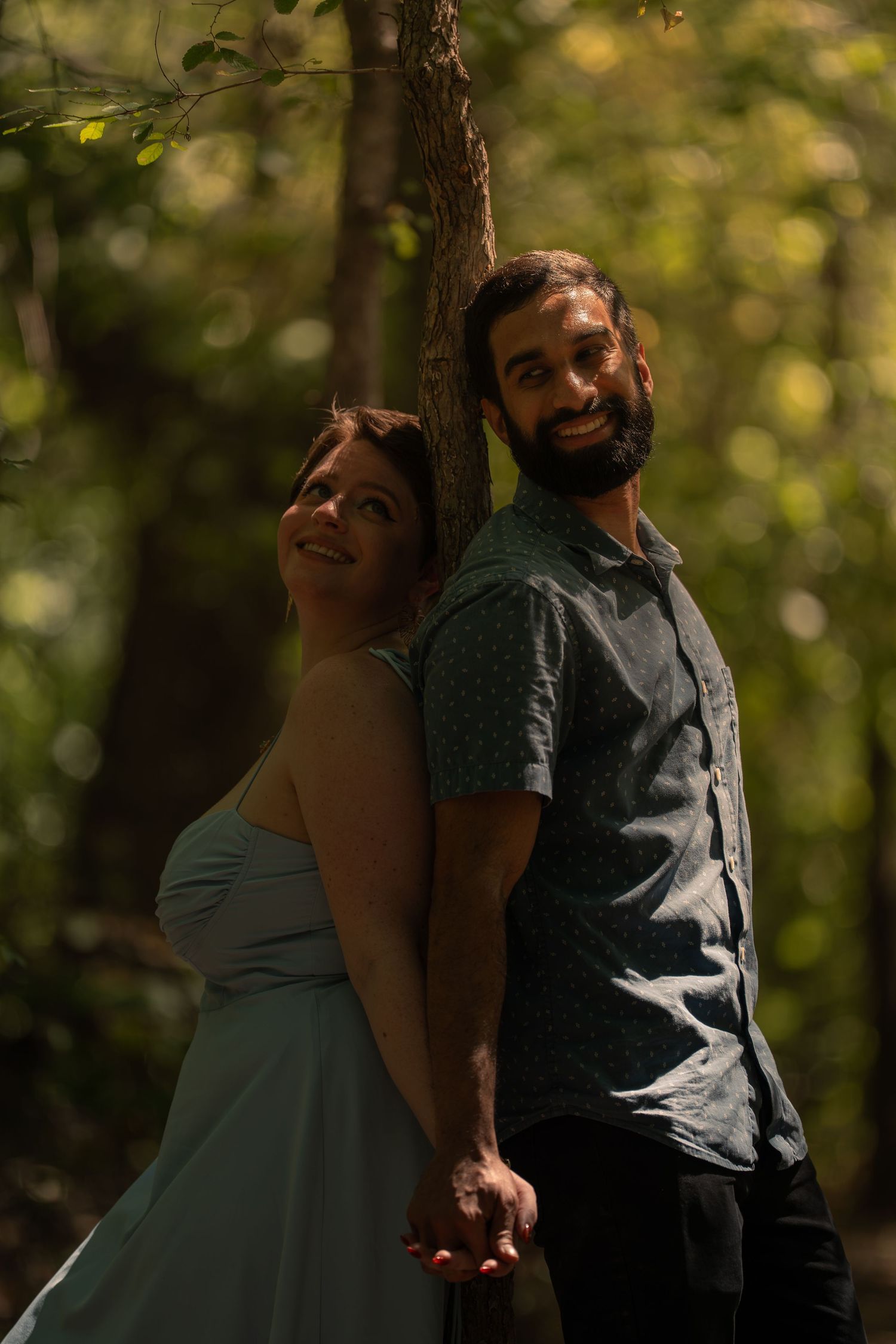 A smiling couple holds hands back-to-back against a tree in a sunlit forest, the woman in white dress, man in gray shirt.