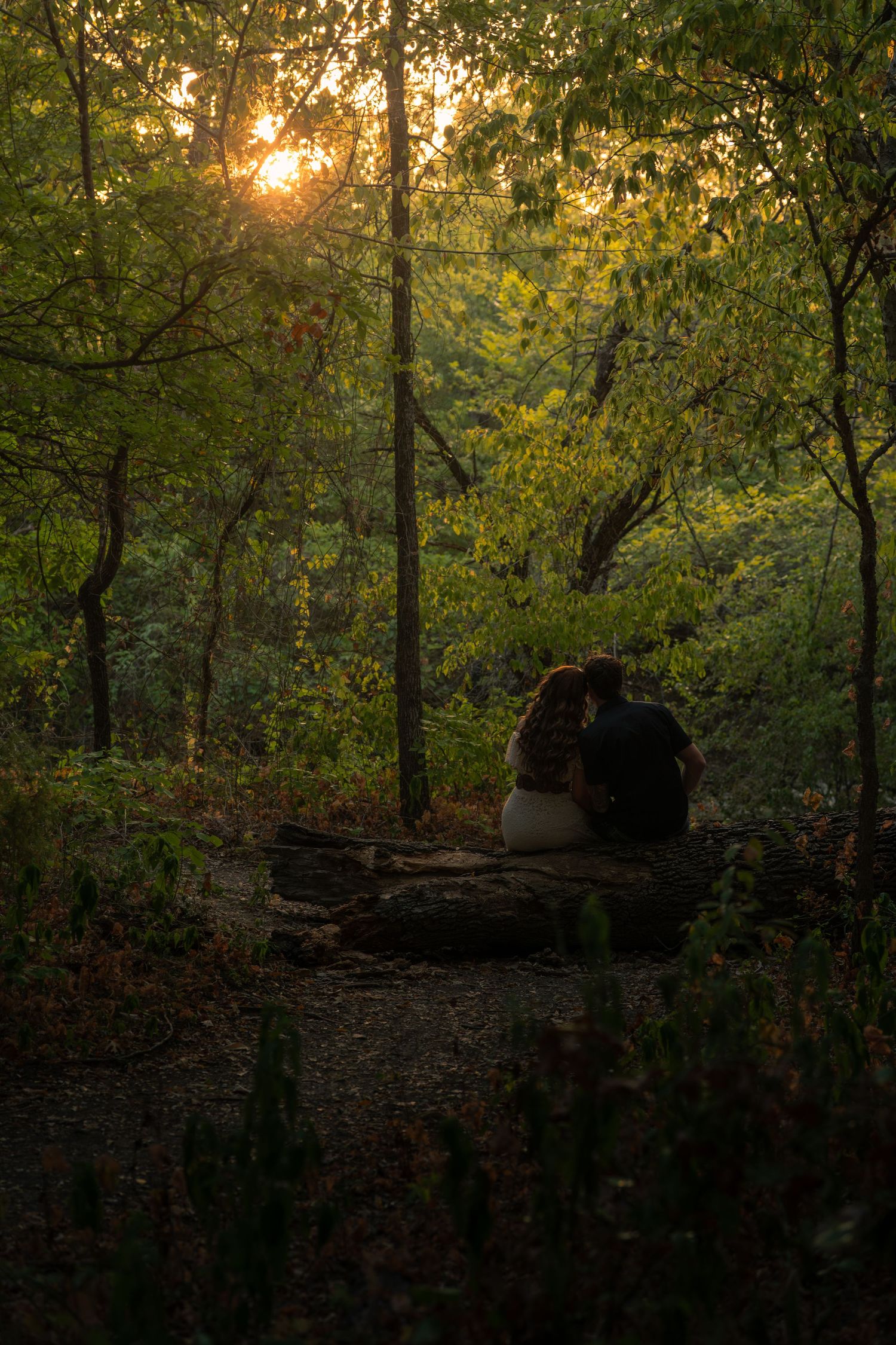 Couple sitting on stone bench at sunset in lush green forest, silhouetted by golden light filtering through trees.