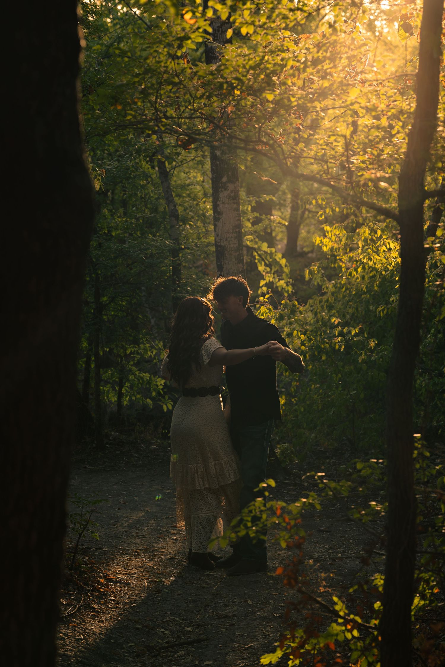 Couple embracing on a sunlit forest path surrounded by lush green trees during golden hour.