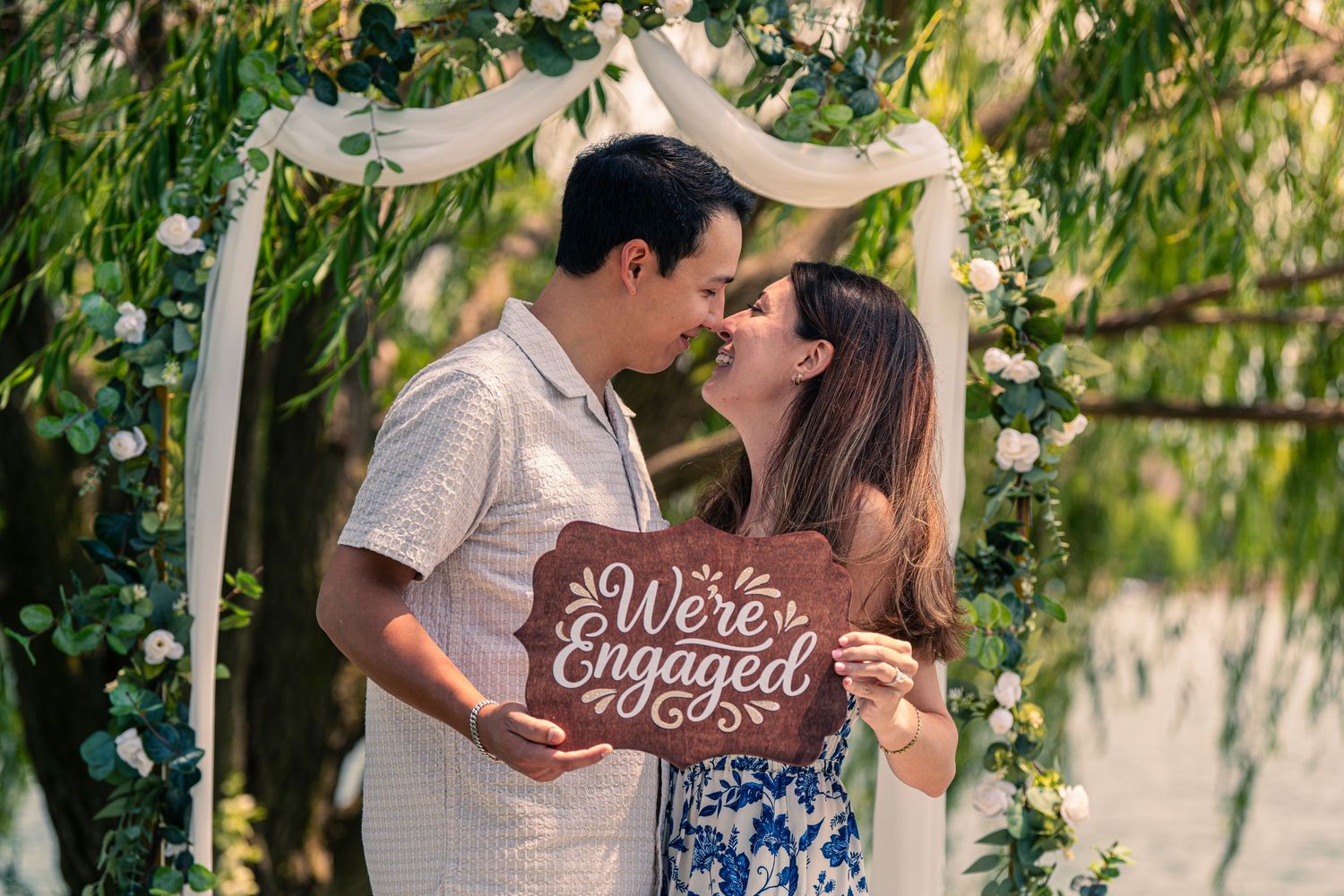 Couple holding We're Engaged sign sharing a kiss under floral arch outdoors.