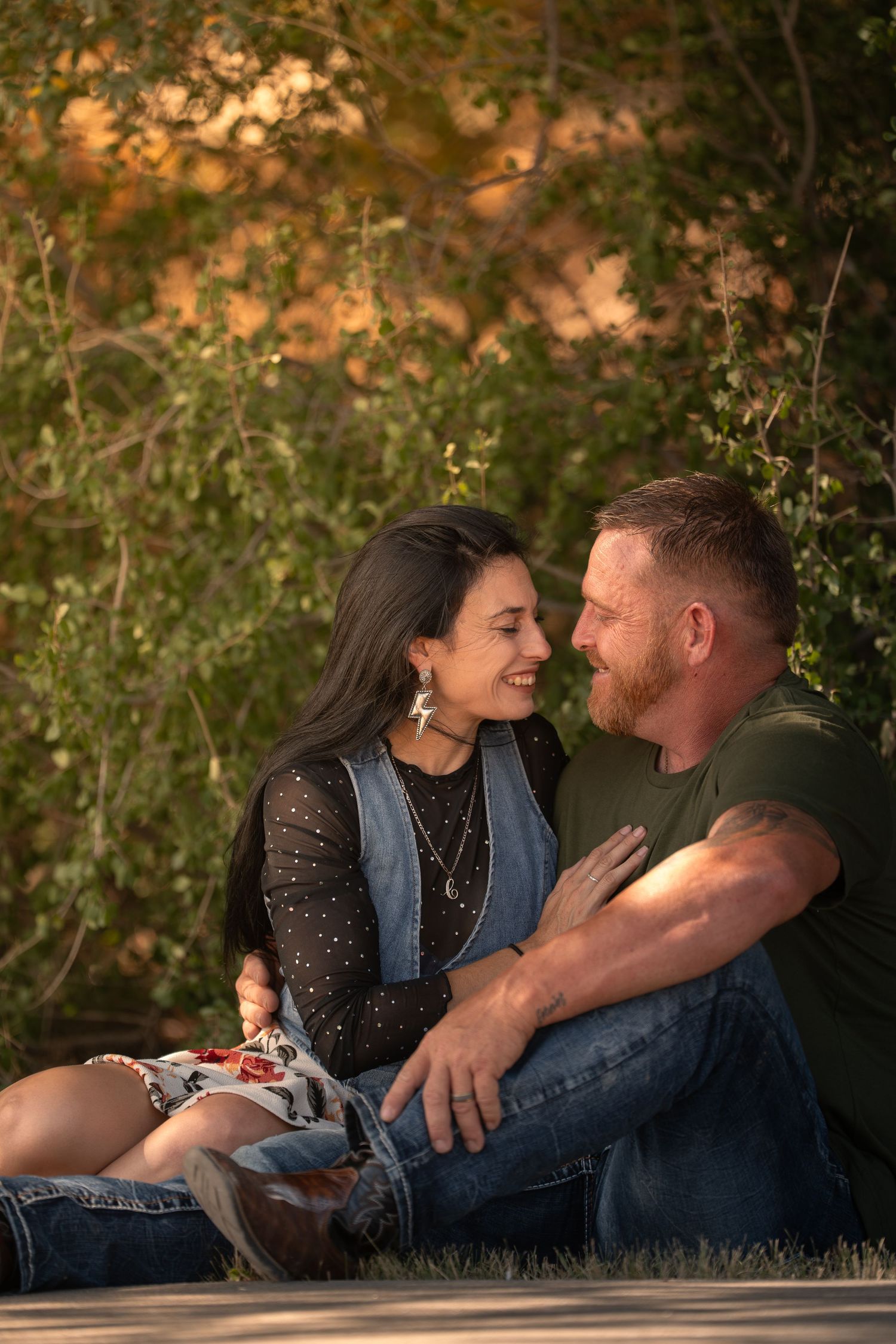 A couple sits closely together on grass, smiling at each other, surrounded by lush green and golden foliage outdoors.