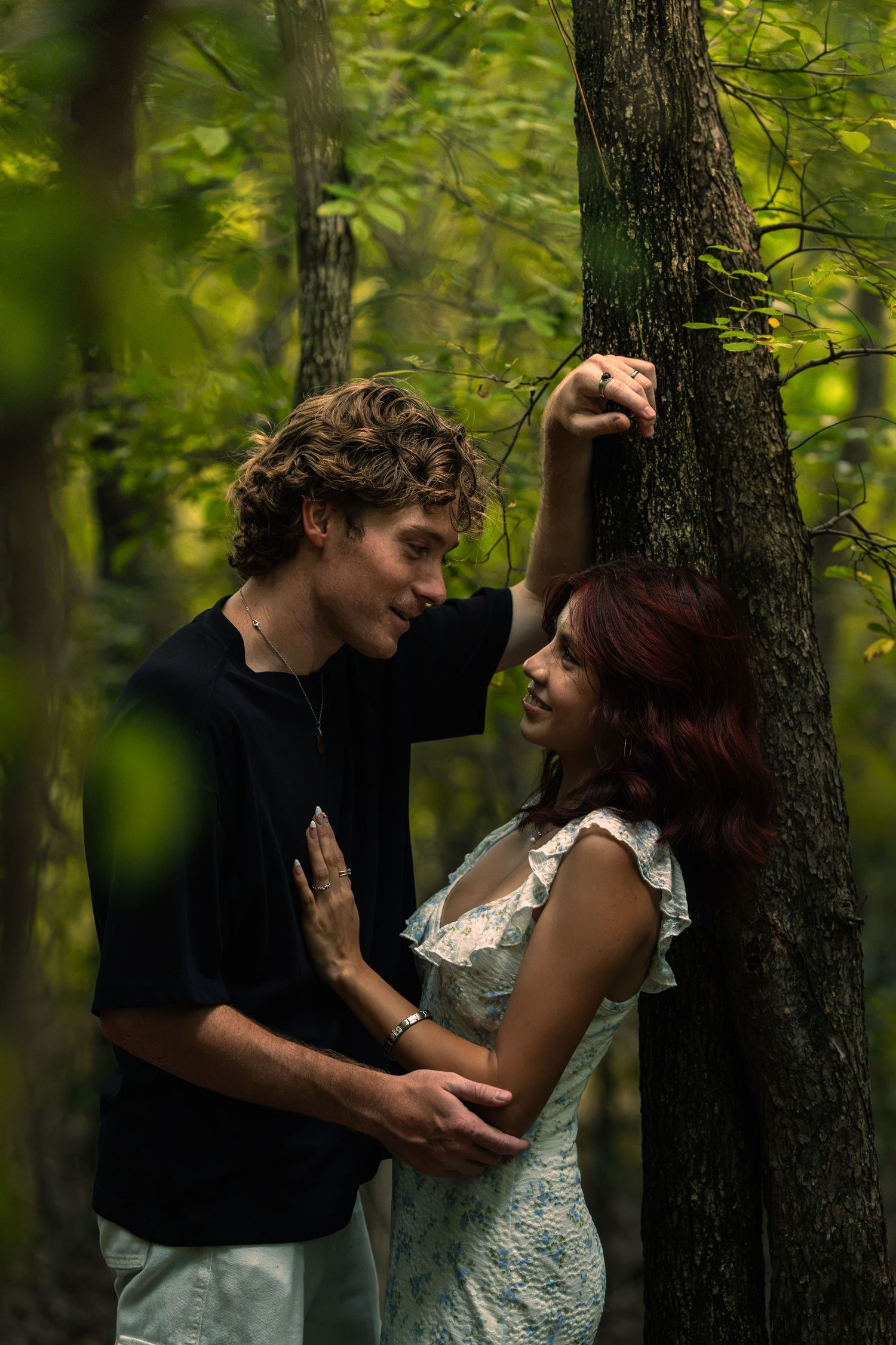 A young couple shares a romantic moment in a lush green forest, the man leaning against a tree toward the woman.
