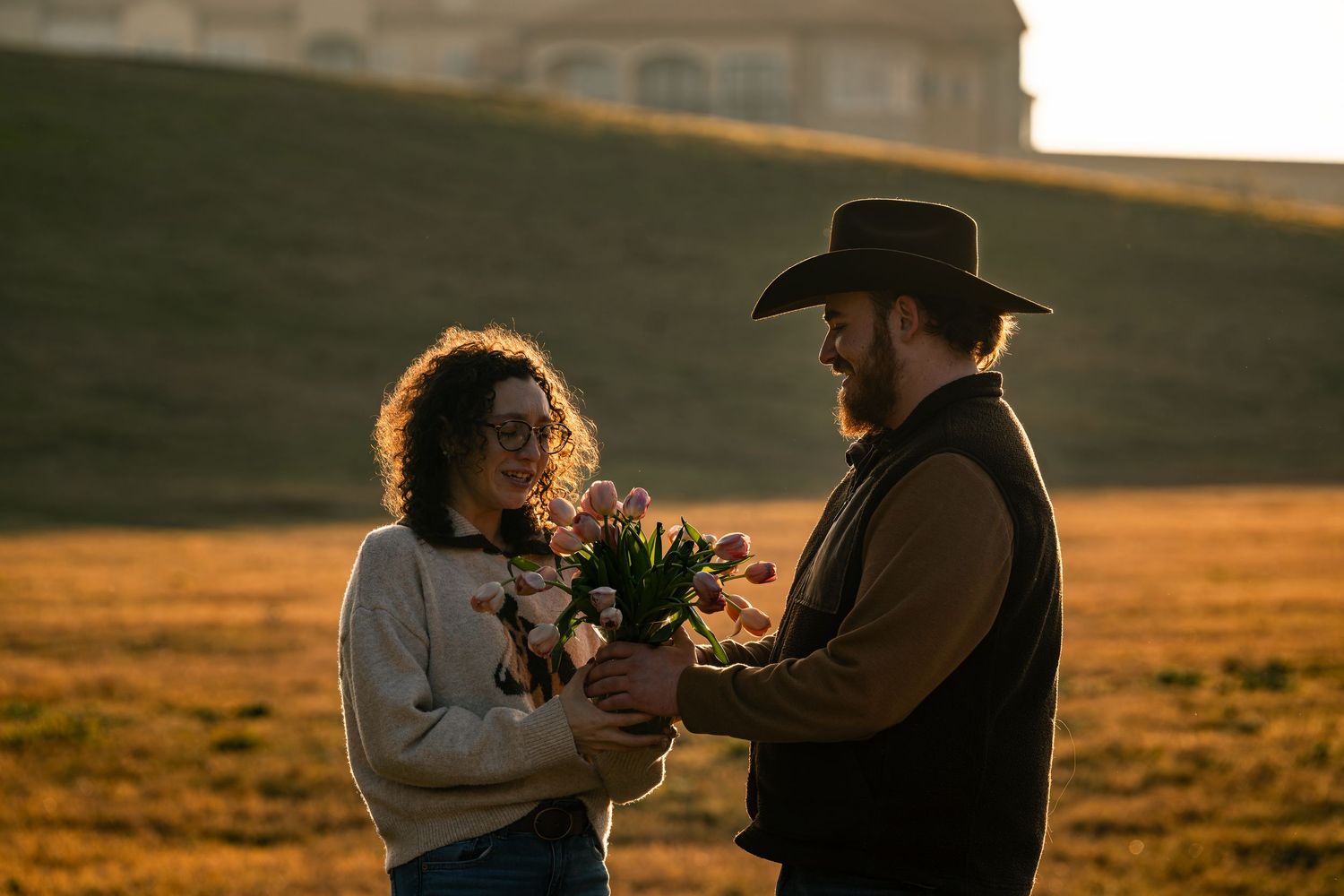 Bearded man in cowboy hat gifting pink tulips to smiling woman at golden sunset.