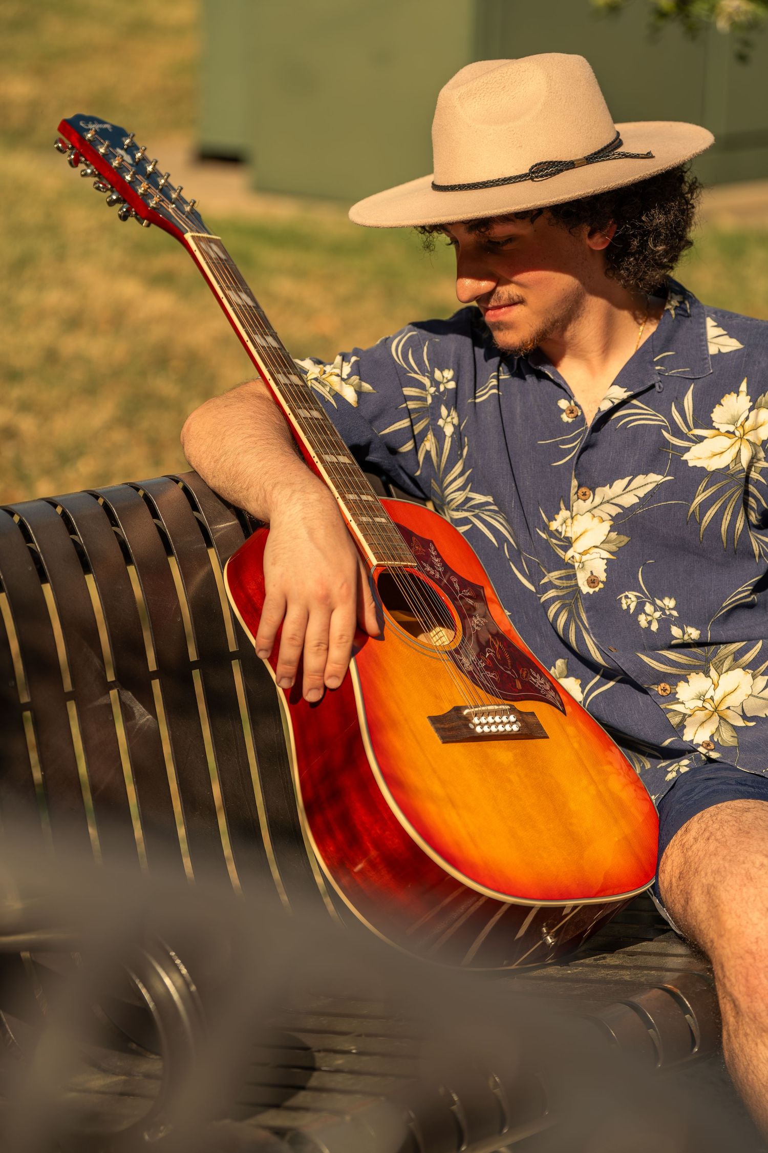 Young man in a floral shirt and hat sitting on a bench holding an acoustic guitar at sunset.