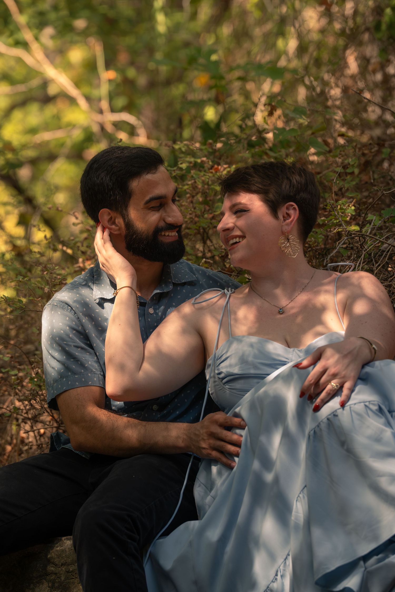 A smiling couple sits together in a sunlit forest, sharing a tender moment in a blue dress and casual shirt.
