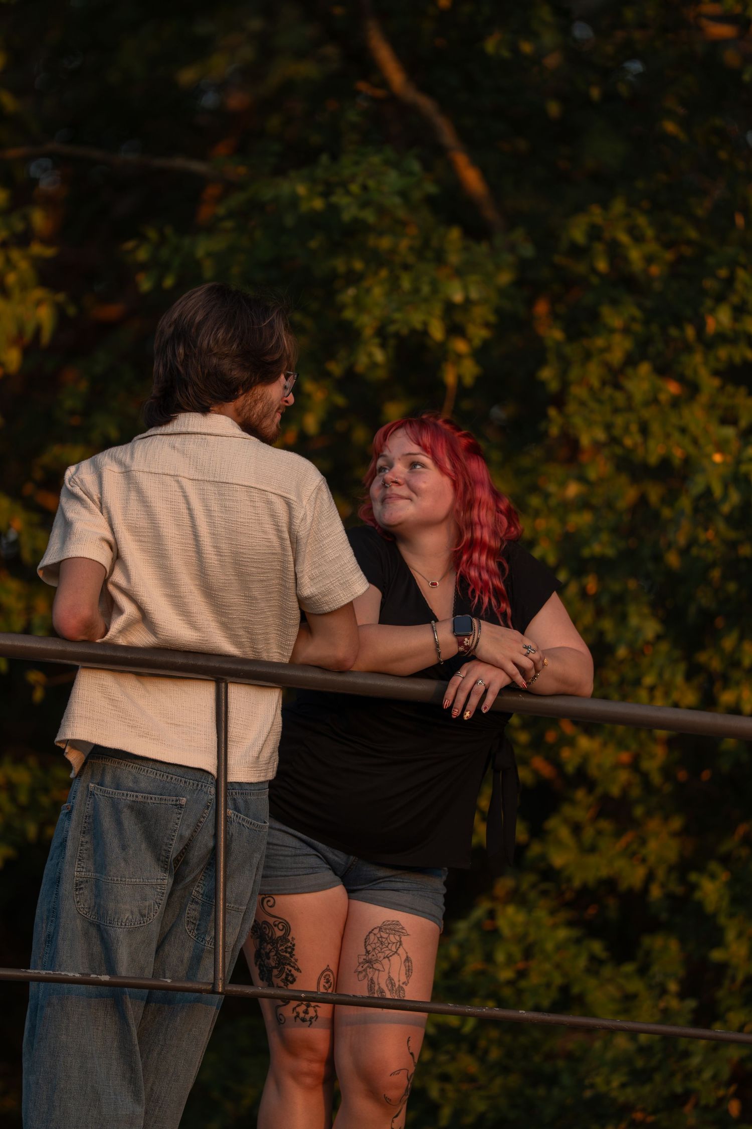 Couple with red-haired tattooed woman in black top leaning on railing, smiling at partner outdoors at golden hour.
