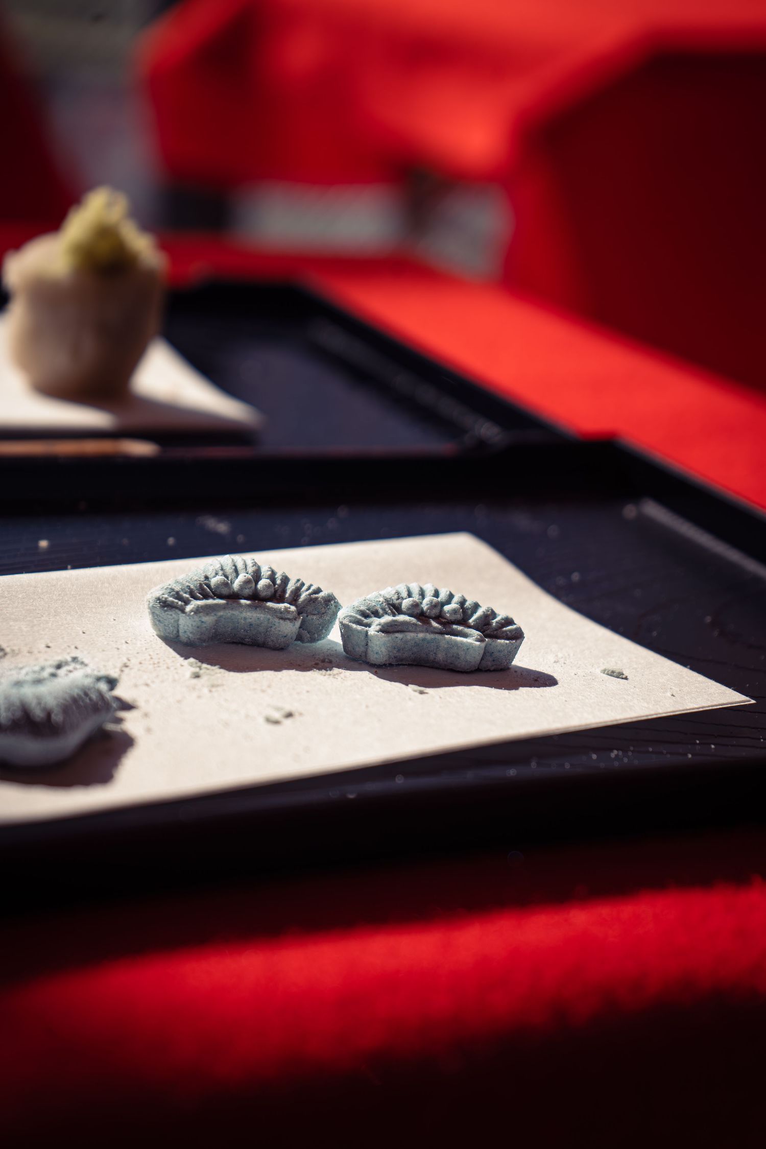 Two gray clay flower-shaped wagashi sweets on white paper atop black Japanese tray with red background.