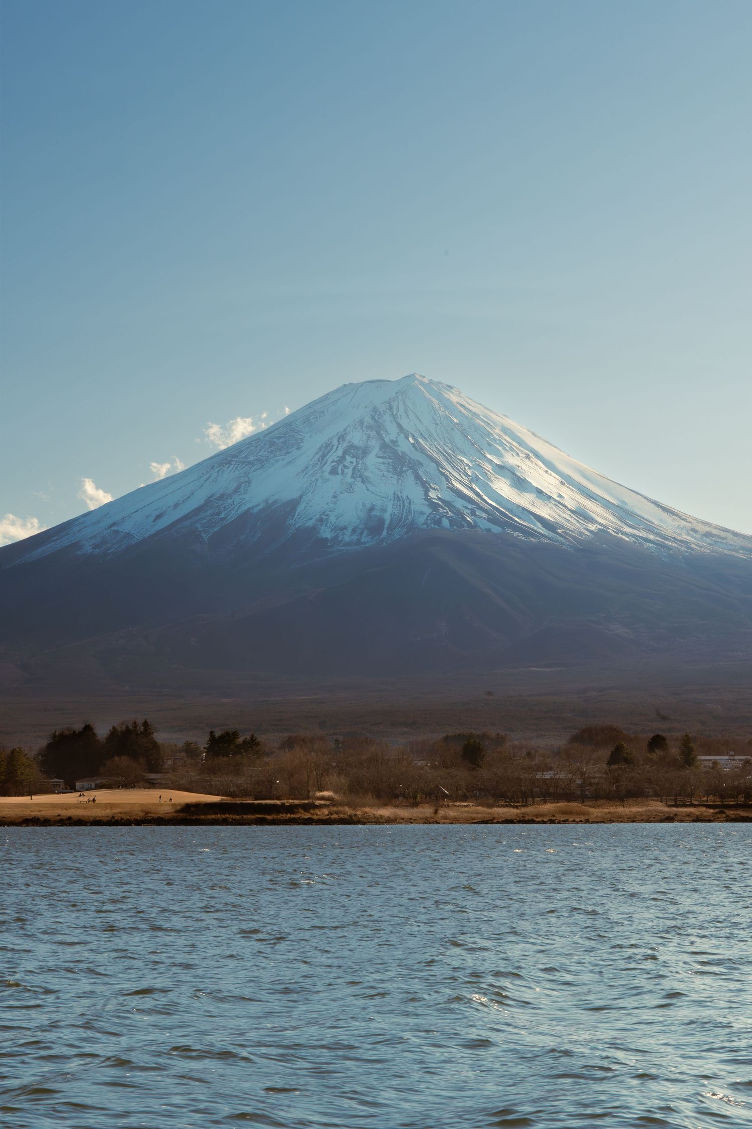 Snow-capped Mount Fuji reflected over calm lake under clear blue sky.