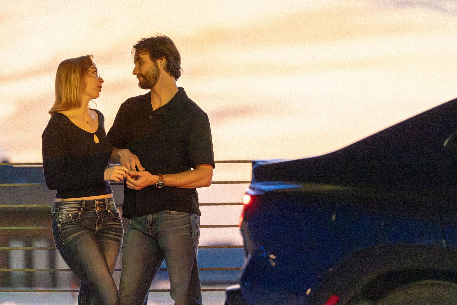 Couple sharing a romantic moment at sunset near a car, woman in jeans and black top, man in black polo shirt.