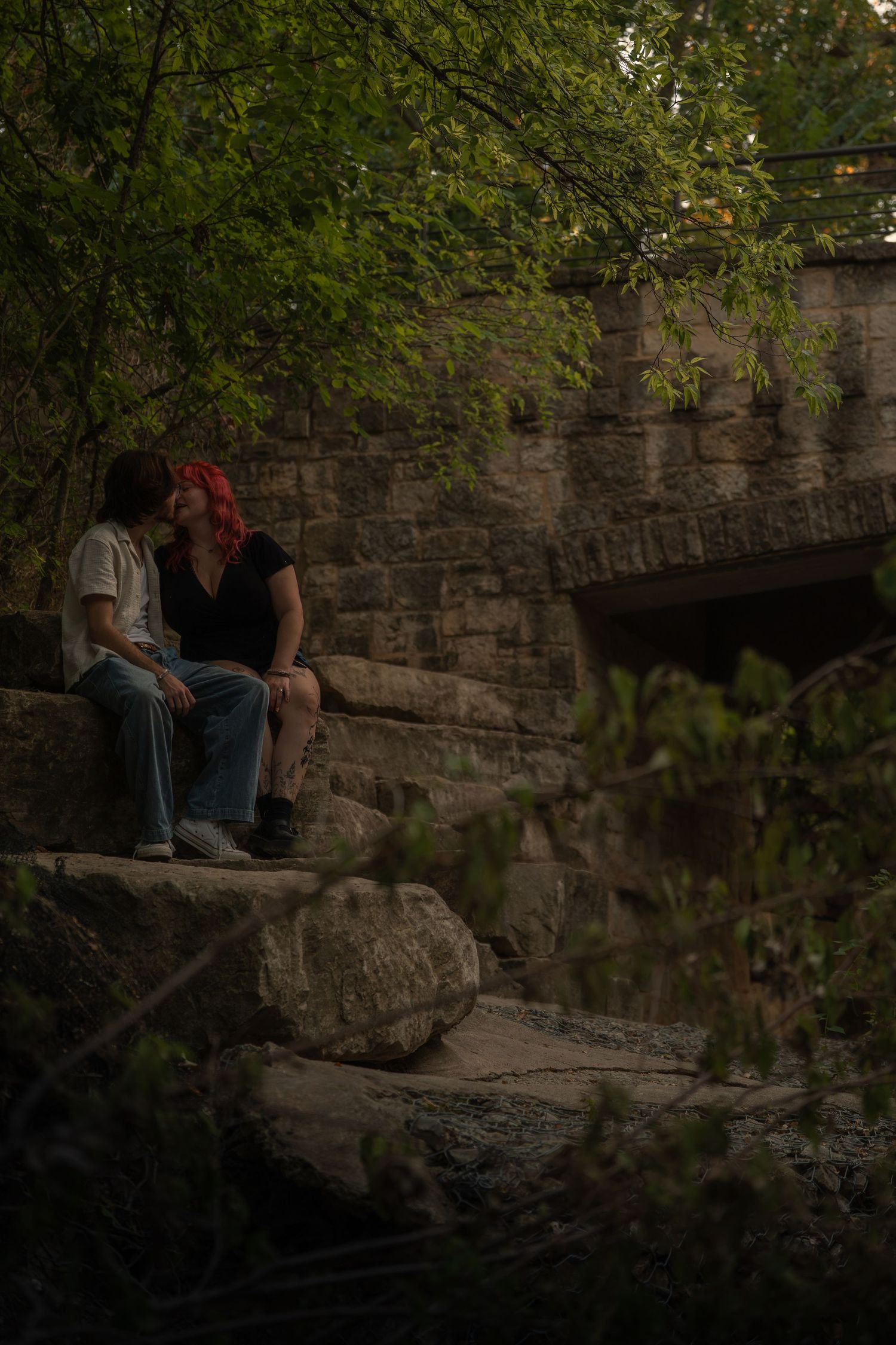 Two people sit closely on rocks near a stone bridge surrounded by lush green trees in a romantic outdoor setting.