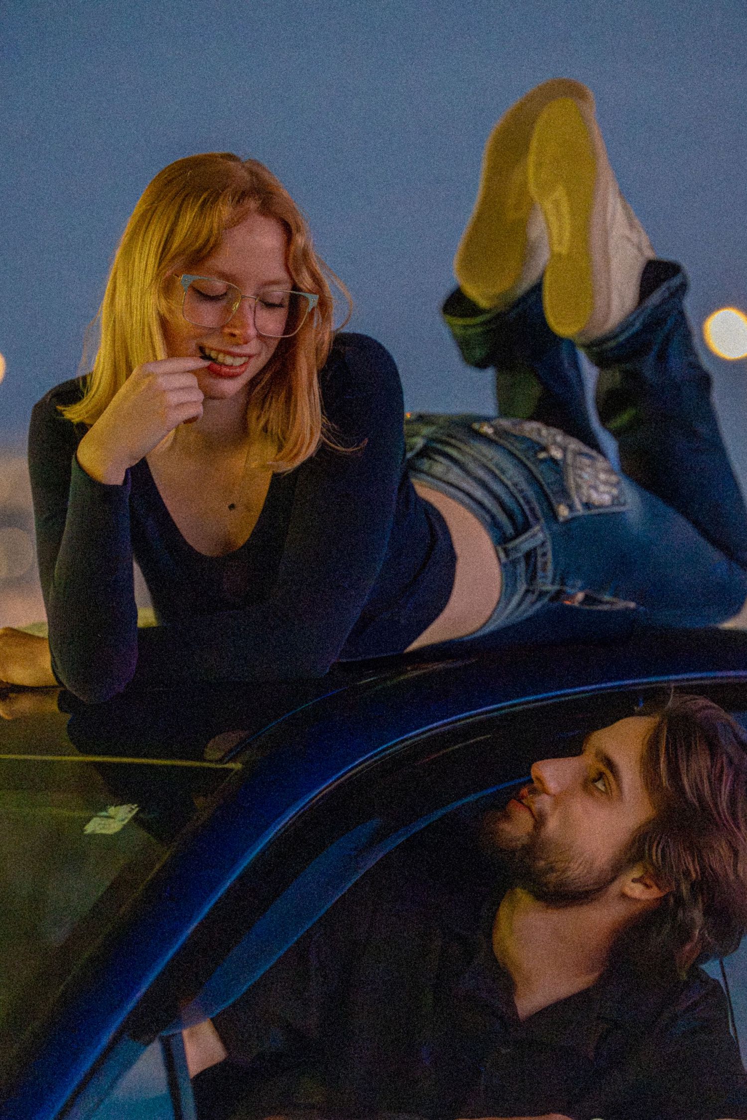 Young woman lying on car roof smiling at man below her during evening outdoor shoot.