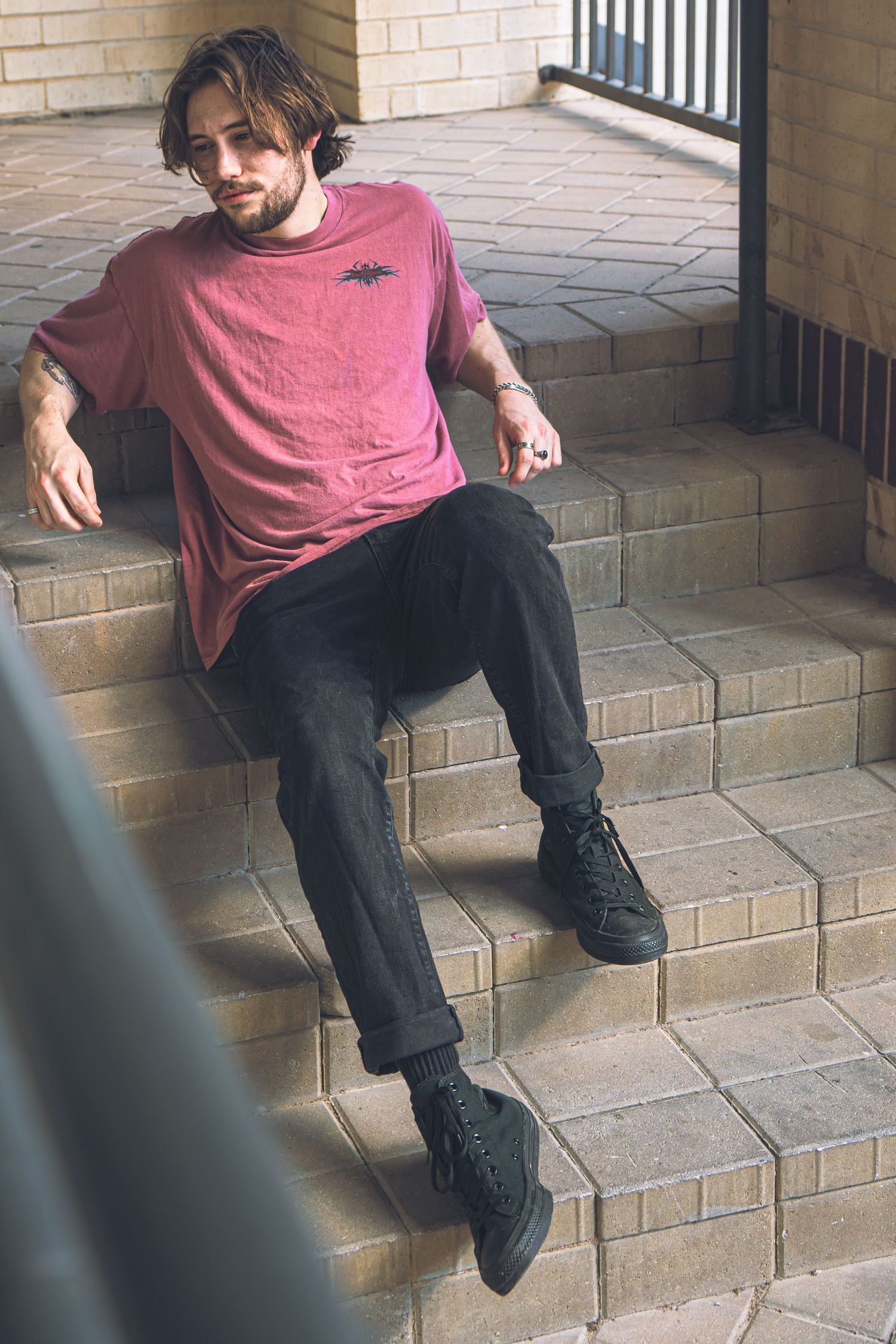 Tattooed man in pink t-shirt and black jeans sitting on brick steps outdoors.