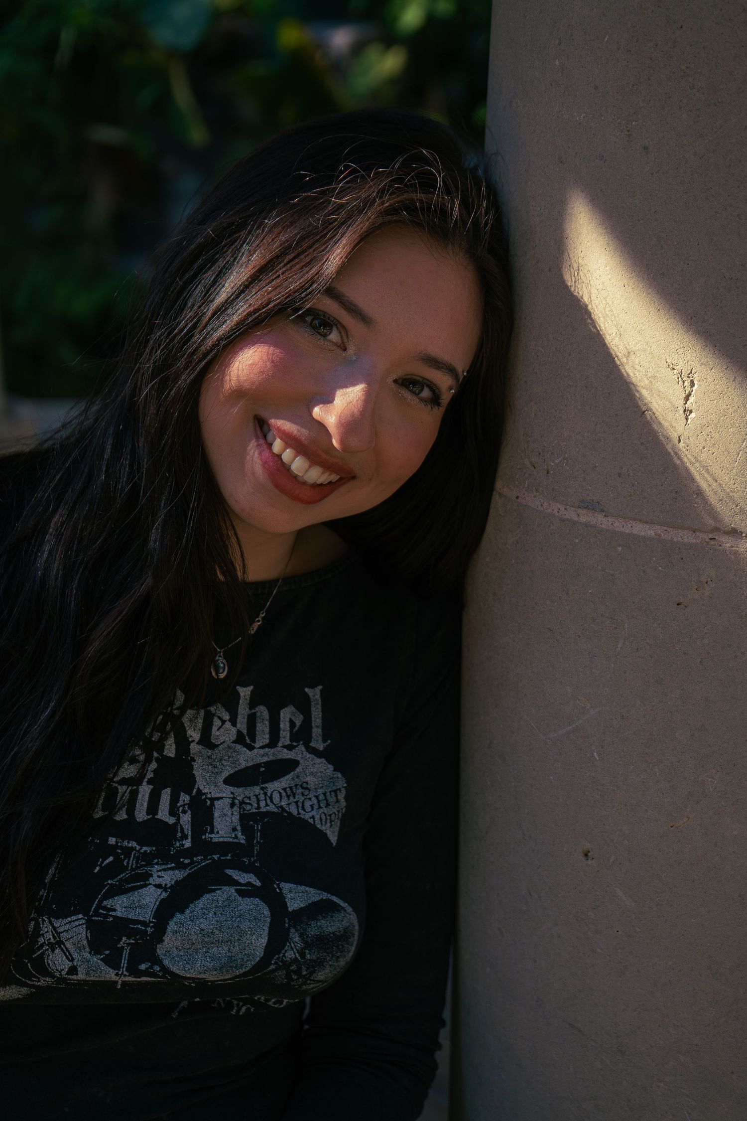 Smiling young woman with dark hair leaning against a stone pillar in sunlight.