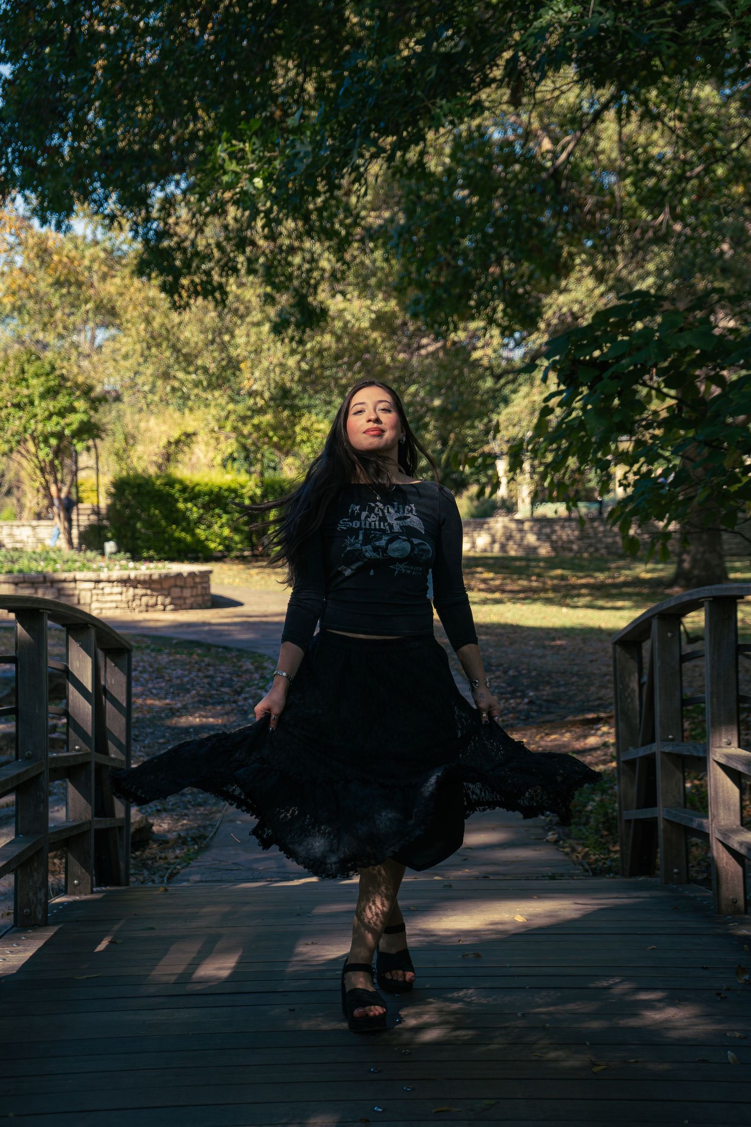 Young woman in black outfit twirling on wooden bridge in sunlit park.