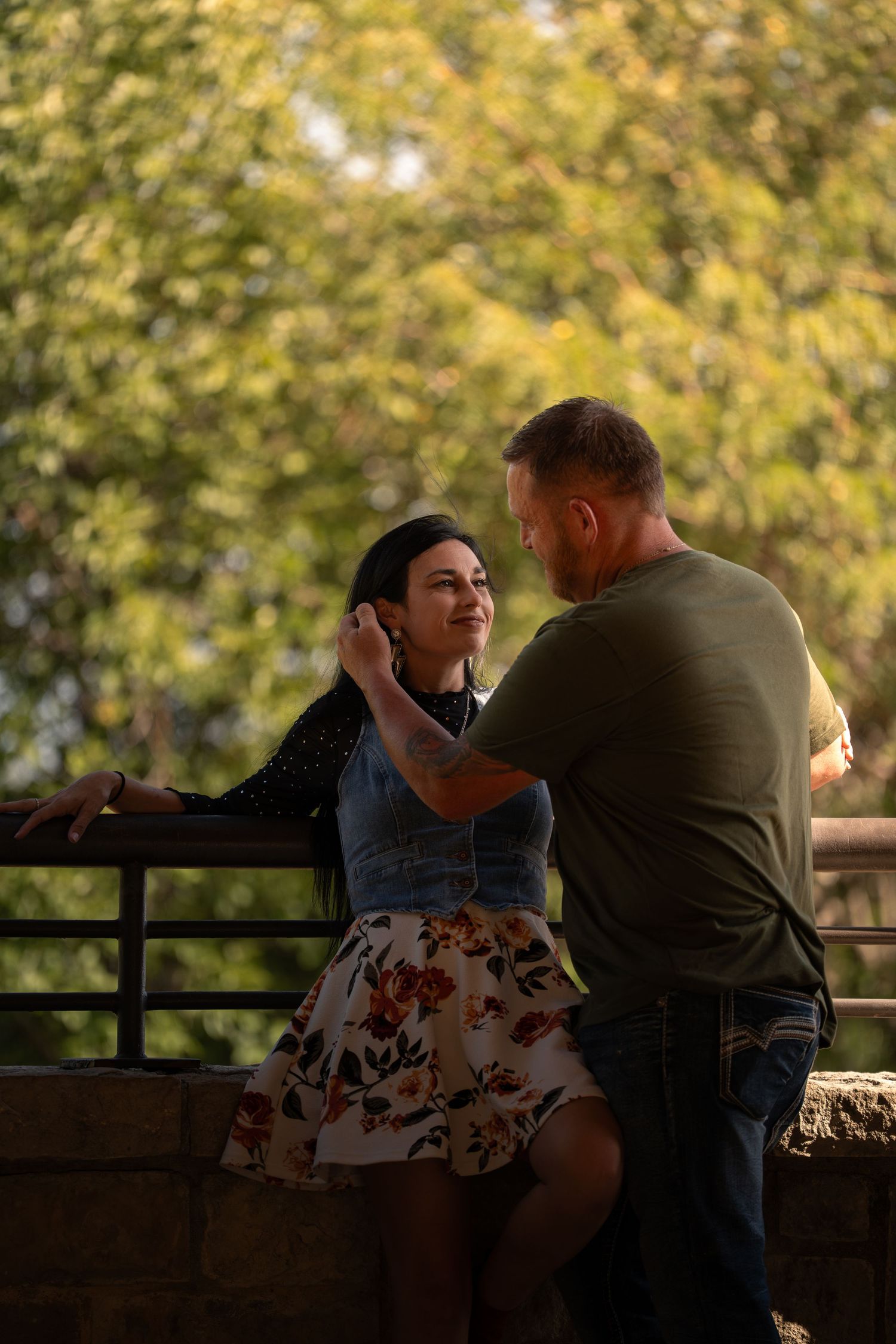 Couple sharing a romantic moment on a bridge surrounded by lush green trees in an outdoor park setting.