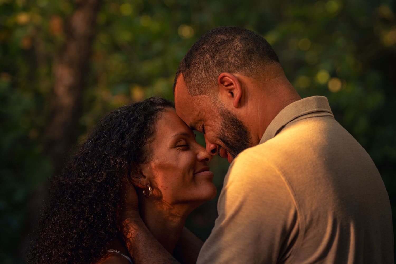 Couple sharing an intimate moment outdoors, foreheads touching in warm golden sunset light with lush green trees behind them.