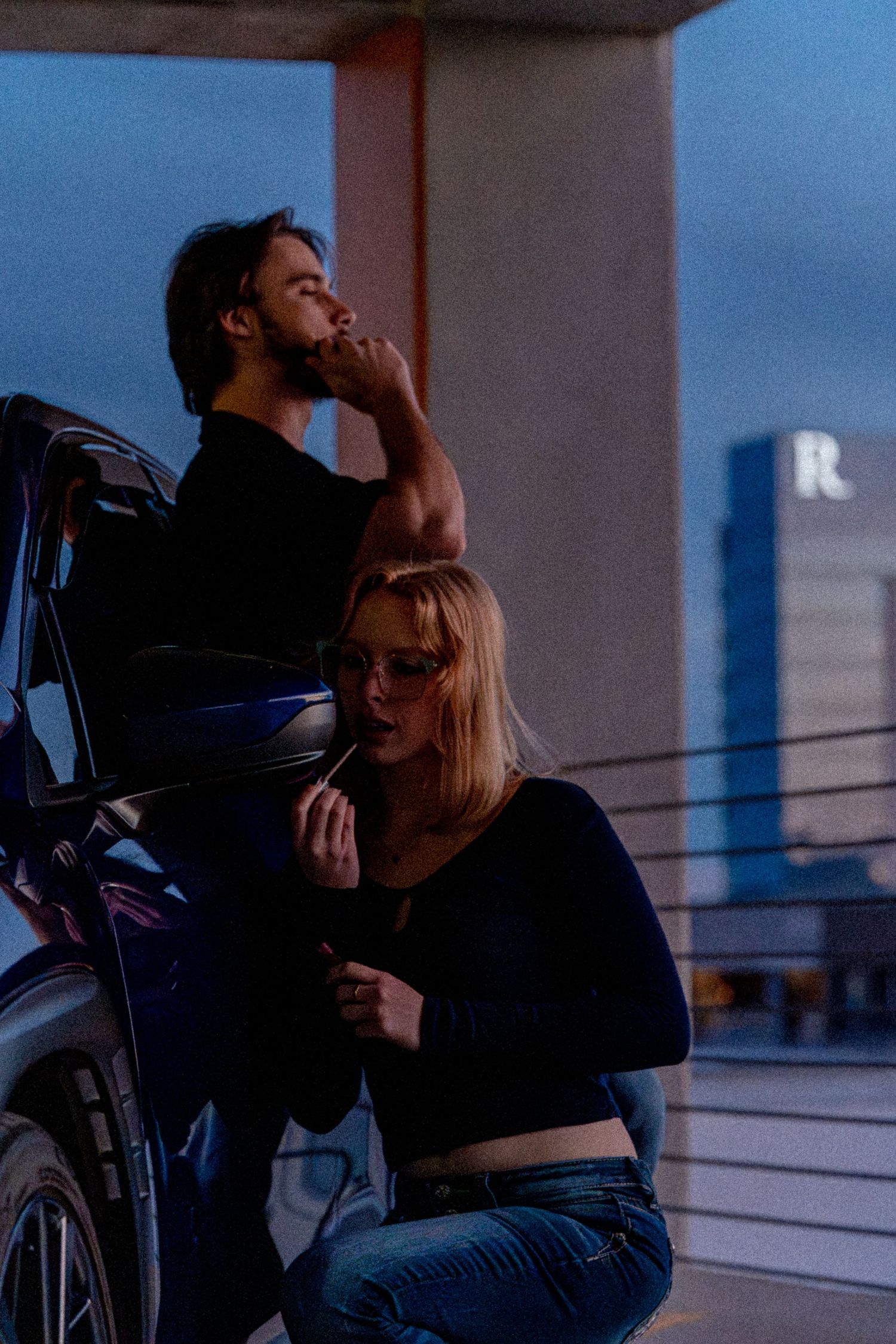 A man and woman in black outfits smoking by a car on a rooftop parking lot at dusk.