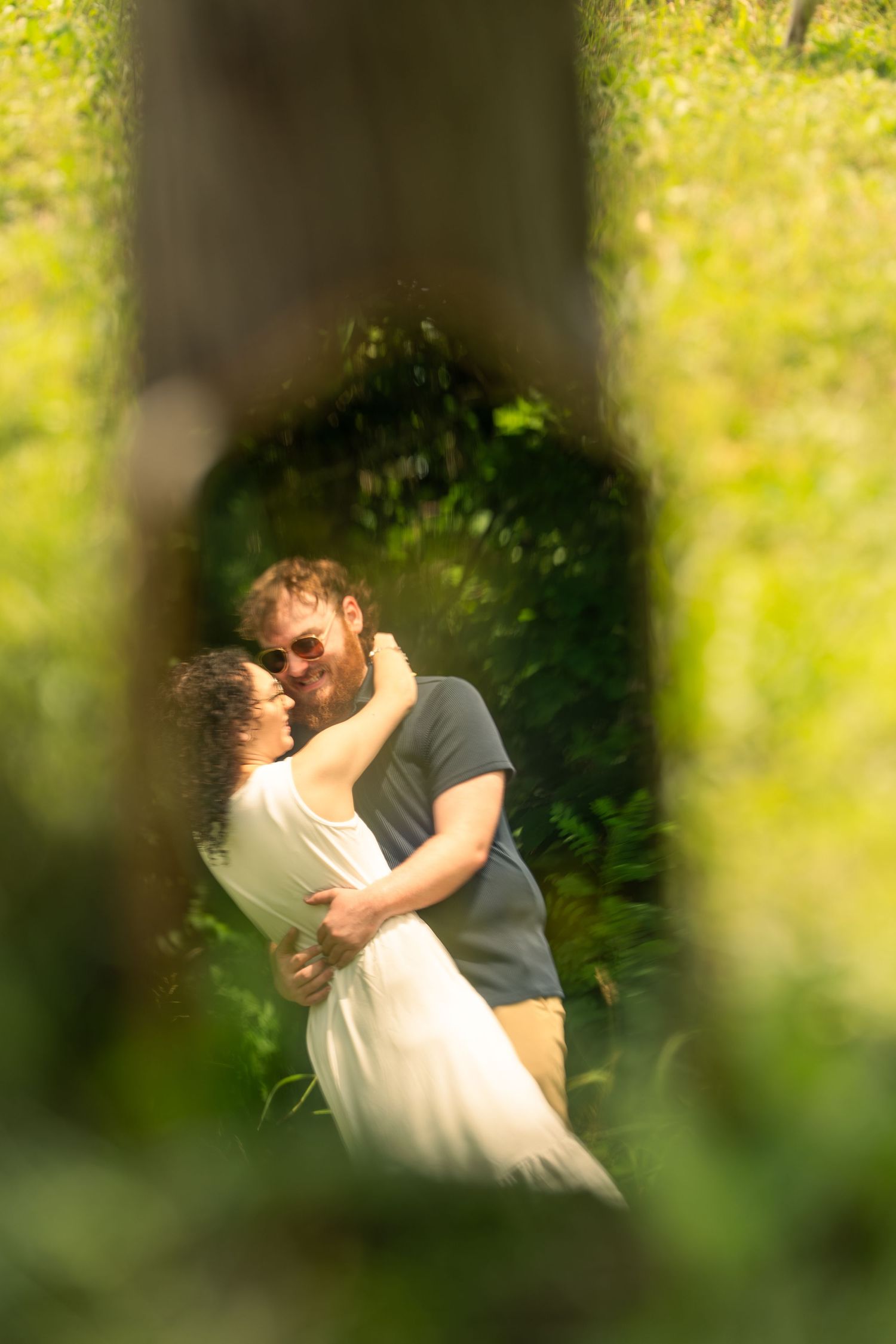 Couple embracing and smiling in lush green garden, viewed through a natural leafy frame.