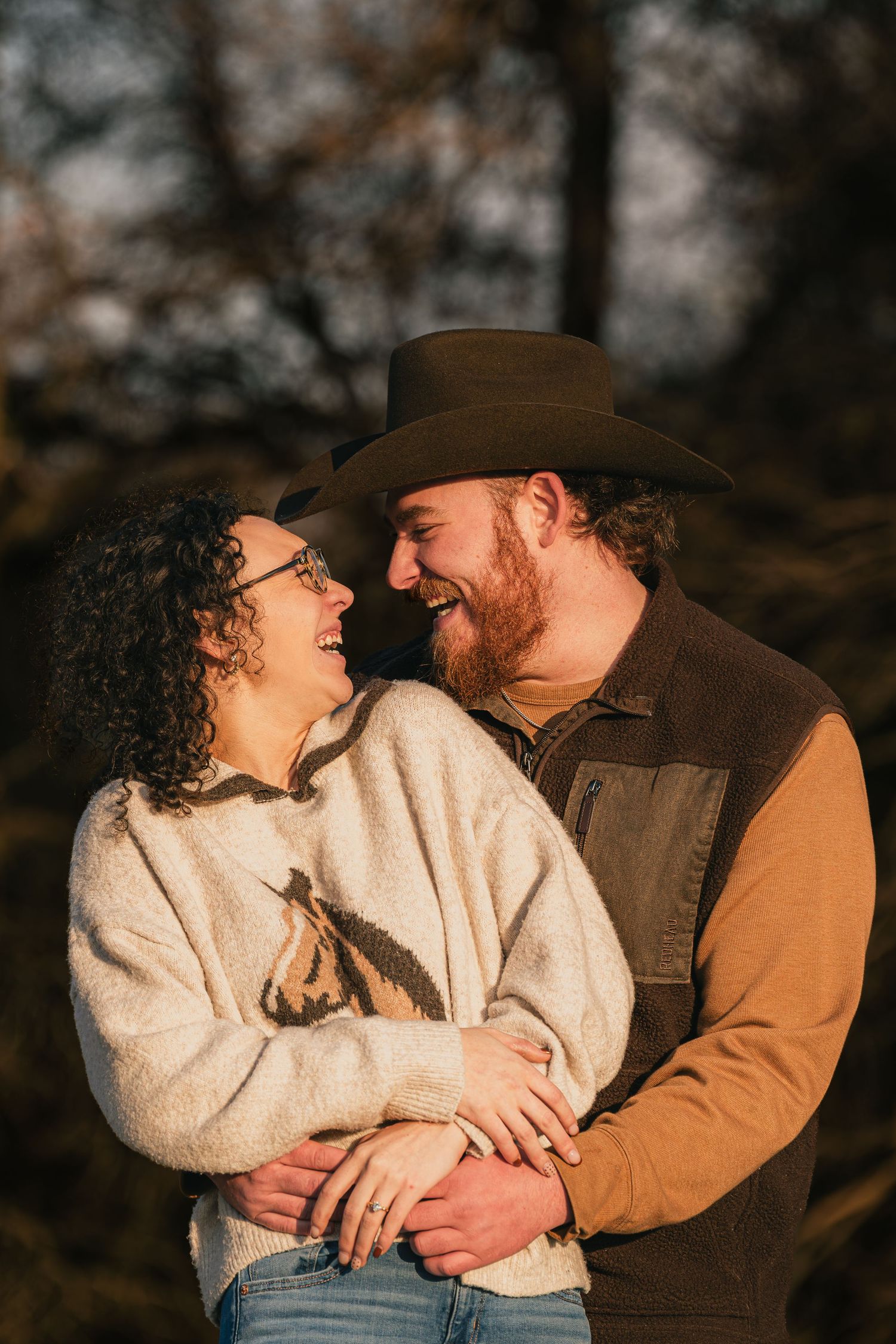 Happy couple laughing and embracing outdoors at golden hour, man wearing brown cowboy hat.
