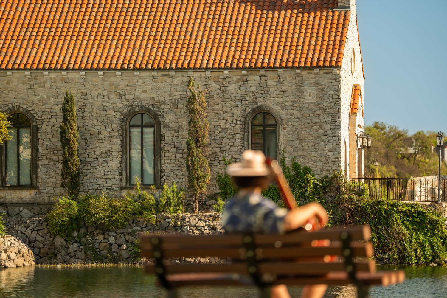 Person sitting on bench overlooking a stone building with terracotta roof and ivy-covered walls.