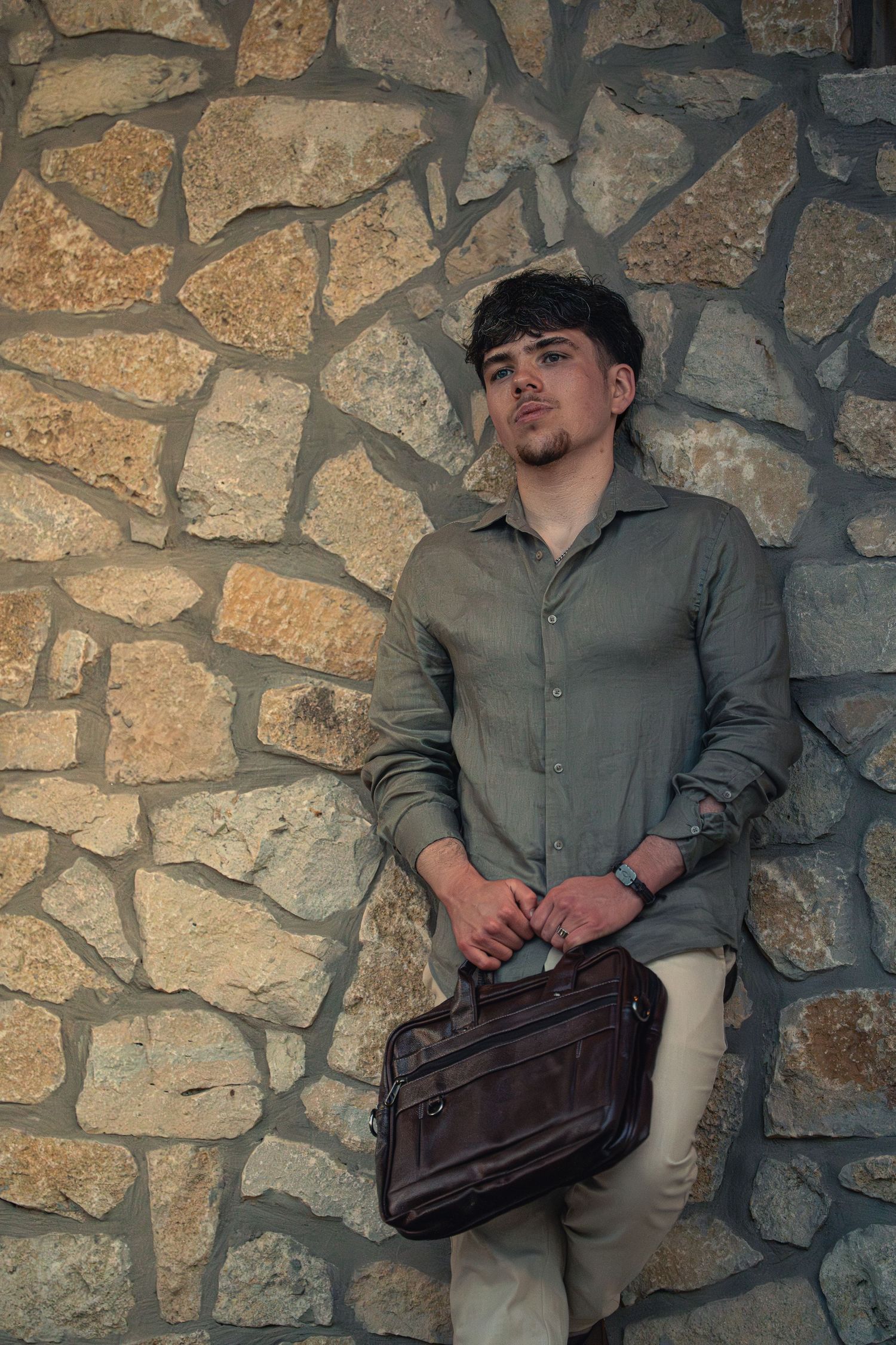 Young man in olive shirt holding leather briefcase, leaning against rustic stone wall.