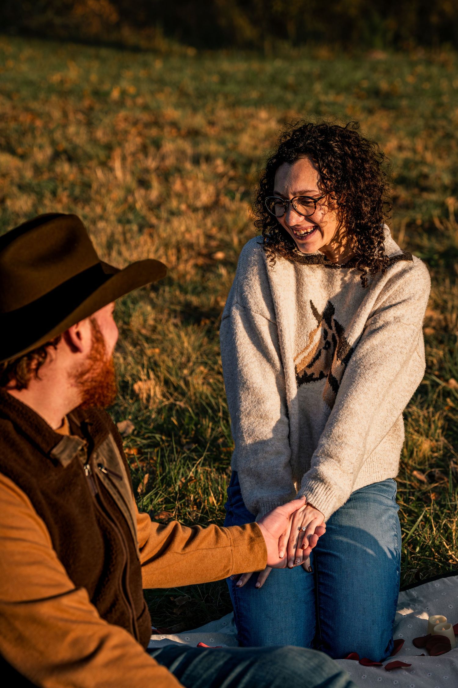Smiling curly-haired woman holding hands with bearded man in cowboy hat on autumn grass.
