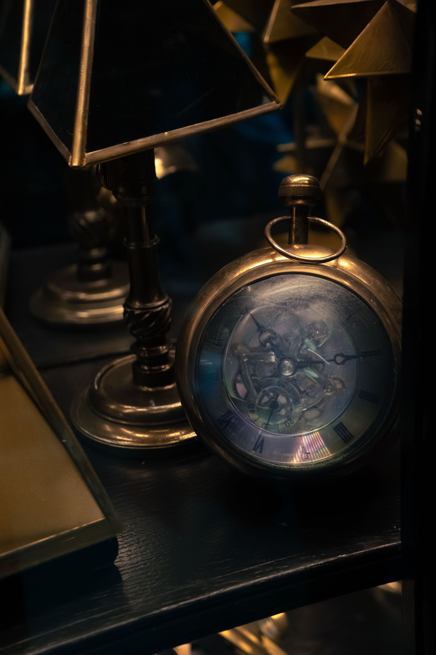 Antique brass skeleton pocket watch with visible gears displayed on dark shelf beside vintage miniature lamp.