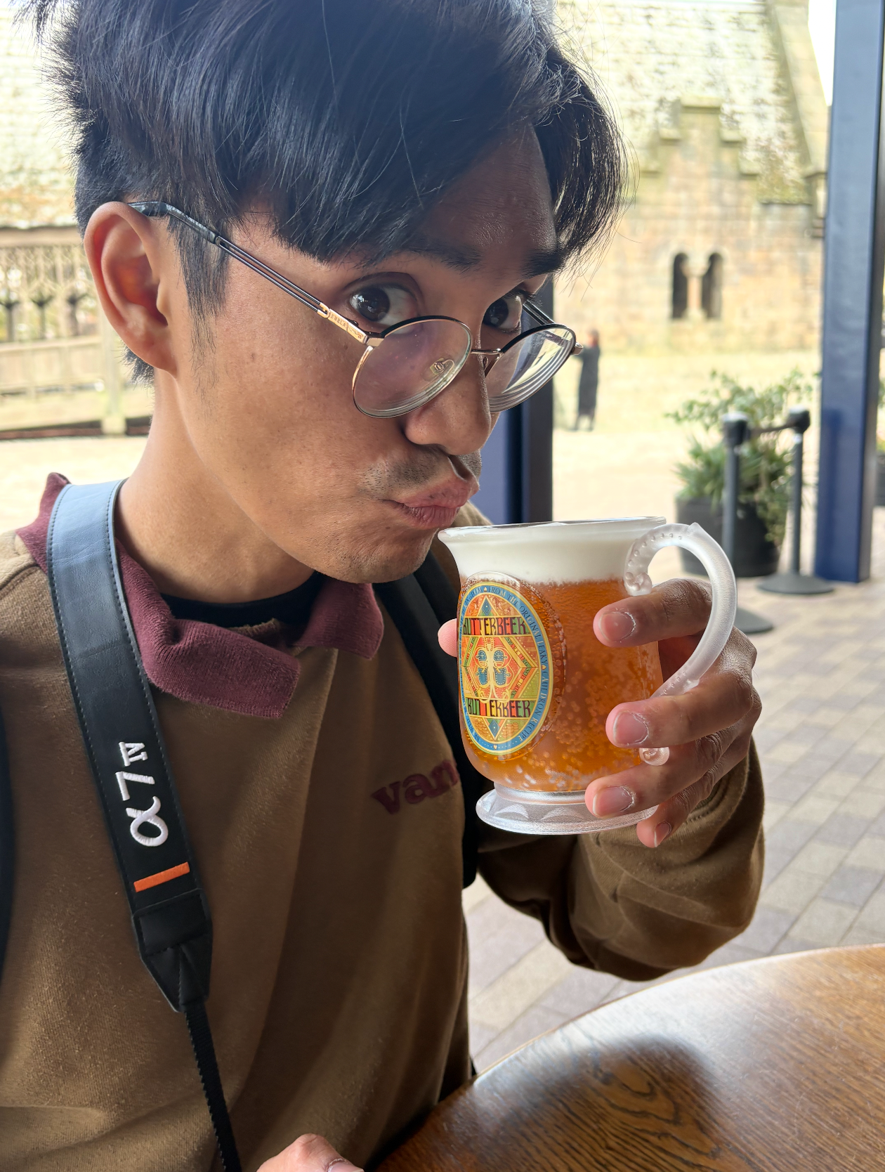 Young man drinking a frothy amber beer from a branded glass mug outdoors at a cafe table.