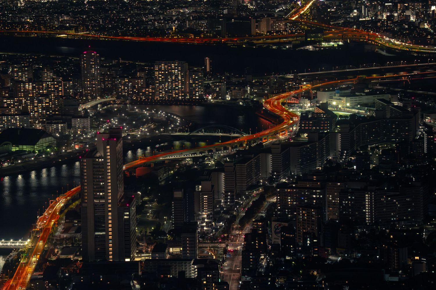 Aerial night view of city skyline with illuminated highways and river reflections.