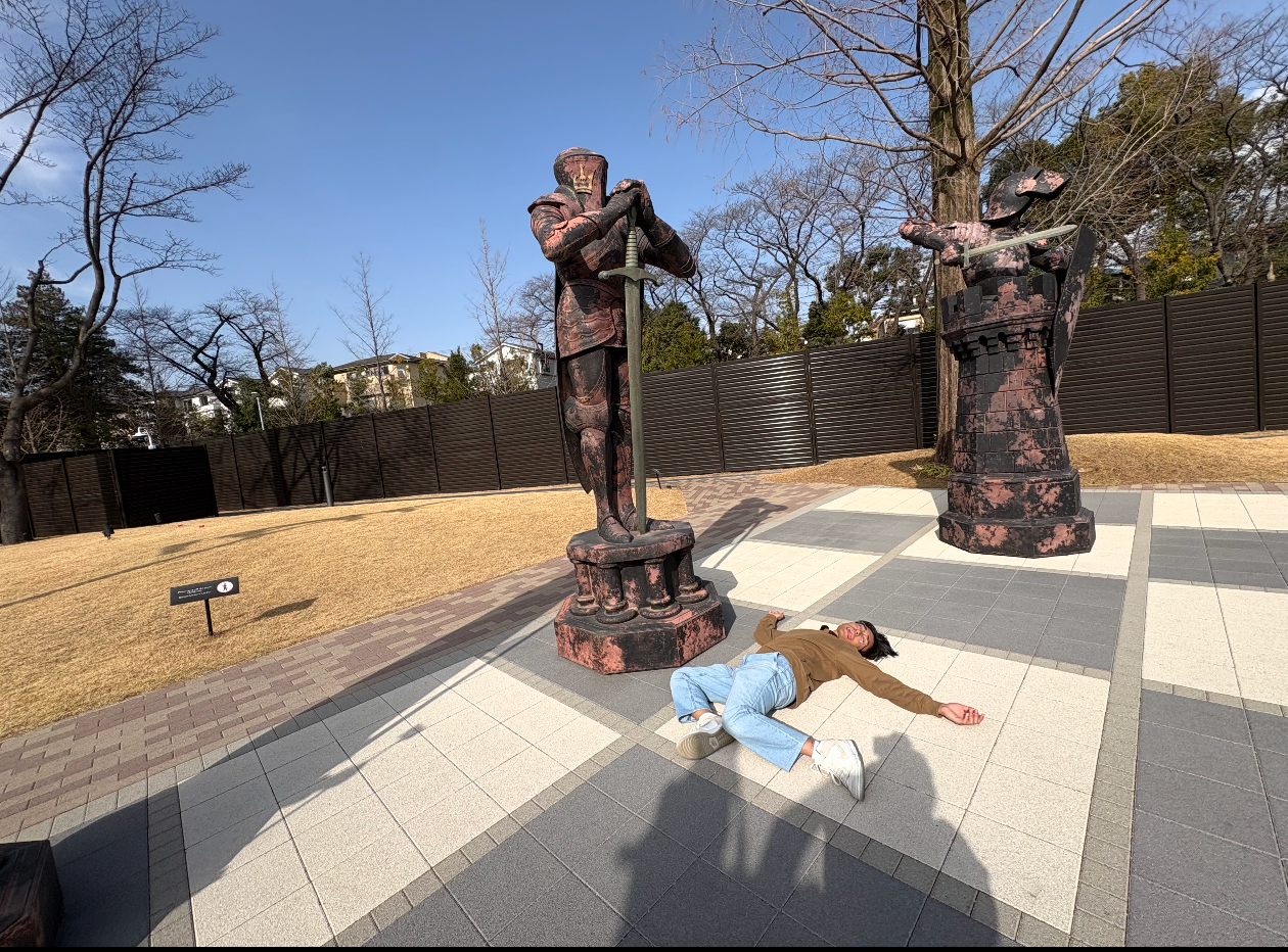 Person lying on the ground between bronze statues in an outdoor sculpture park on a sunny day.
