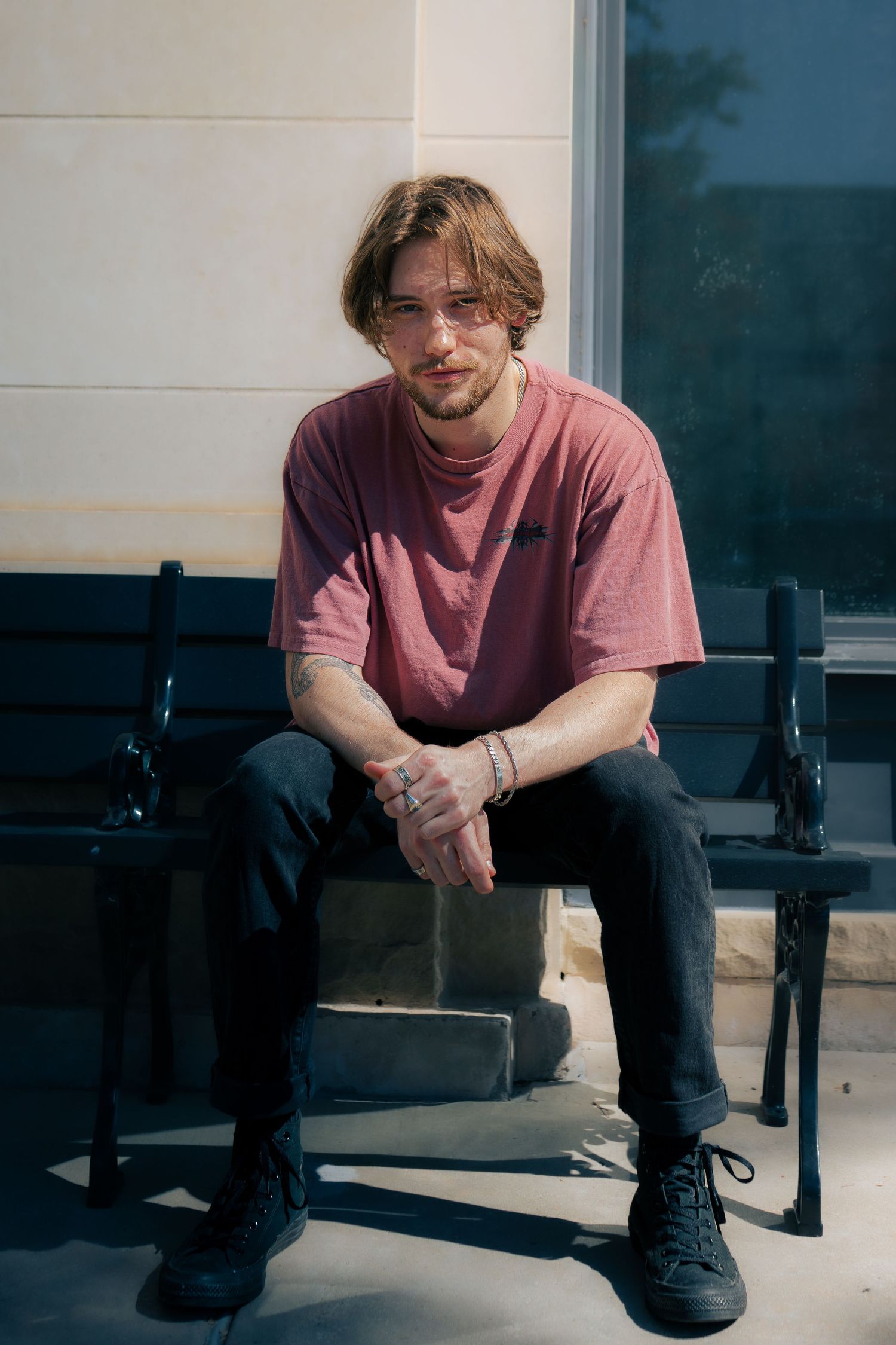Young man in pink t-shirt and black jeans sitting on a bench in sunlight.