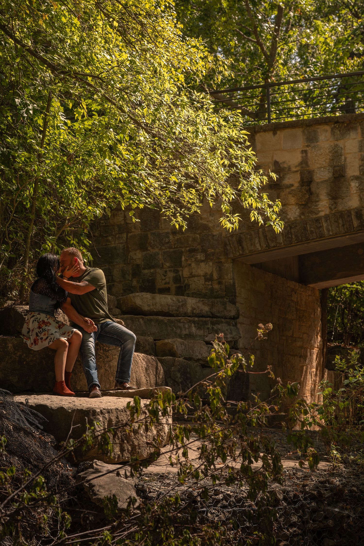 Couple sitting on rocks near a bridge surrounded by lush green trees in a romantic outdoor setting.