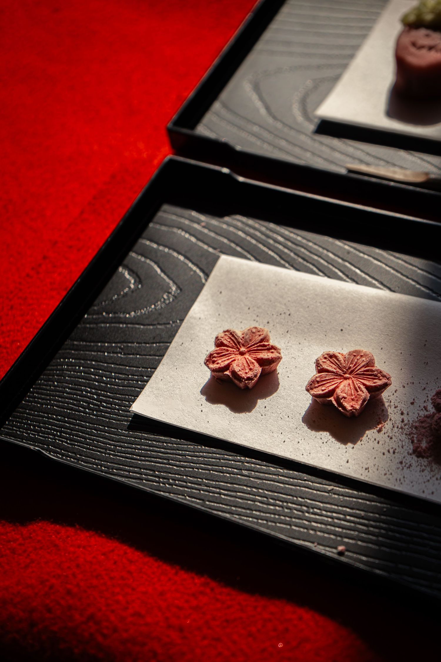 Two red sakura flower-shaped Japanese wagashi sweets on white paper displayed on black wooden tray.