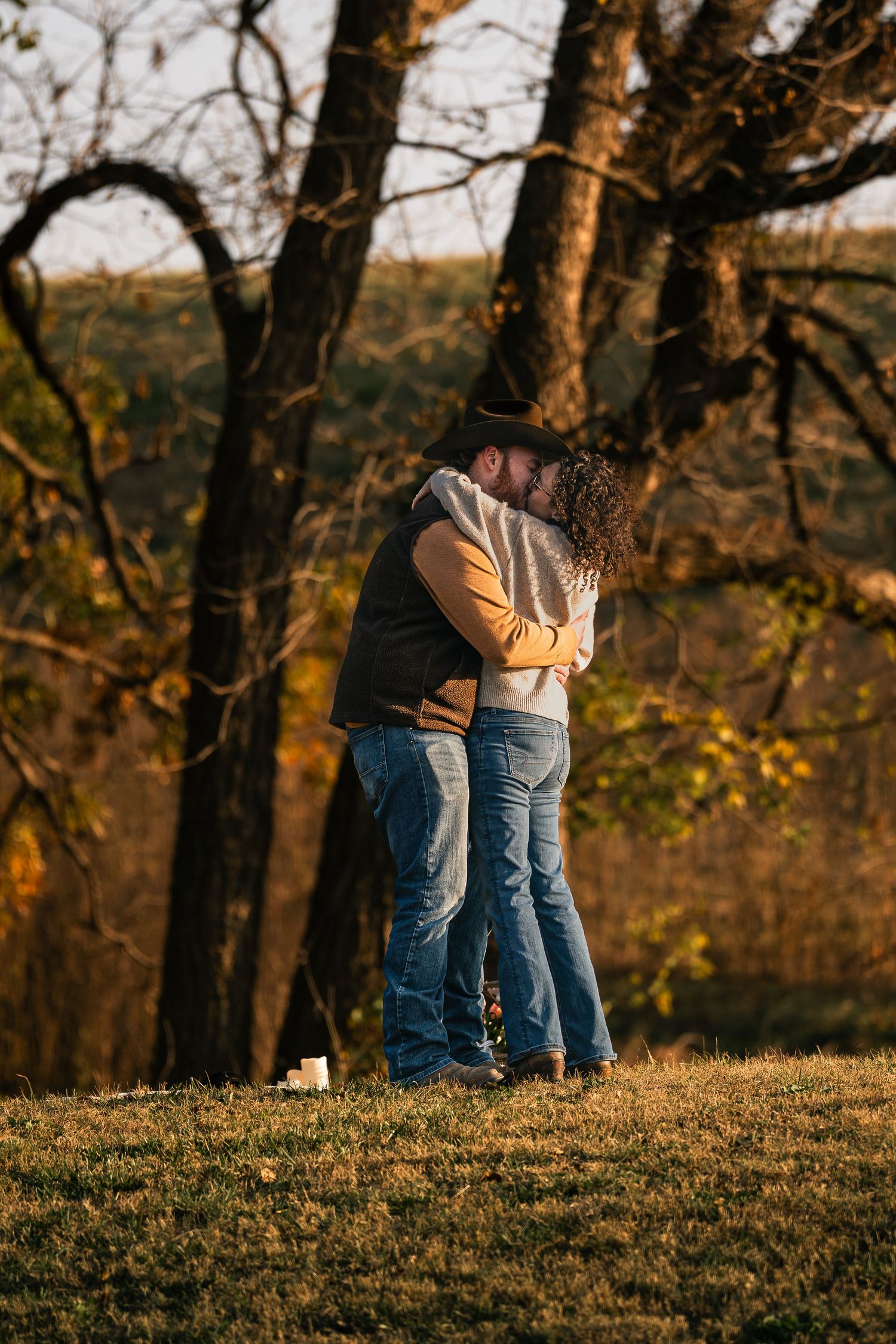 Romantic couple kissing outdoors during golden hour with colorful fall foliage in background.