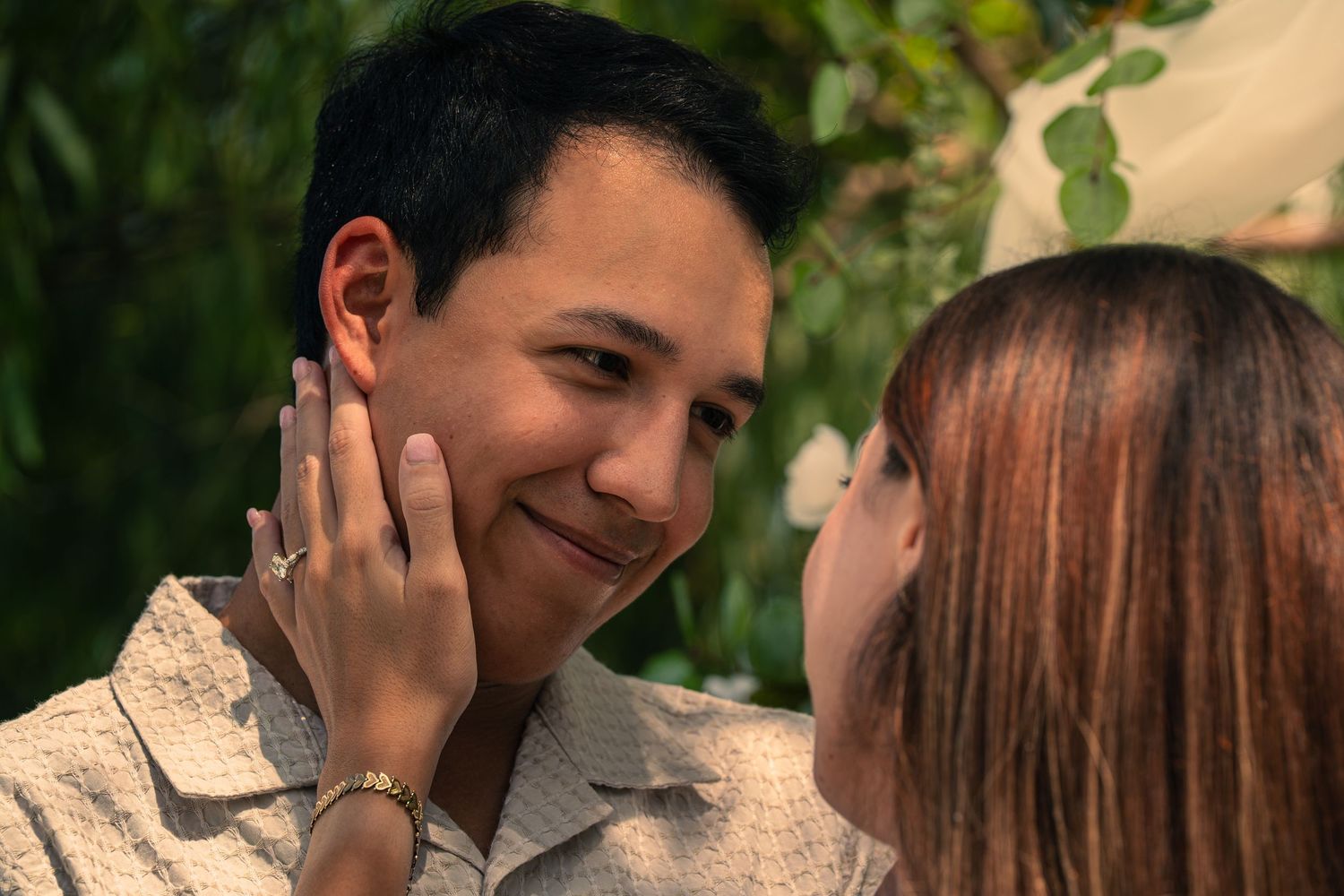A woman tenderly touches a smiling man's face in a romantic outdoor moment surrounded by greenery.