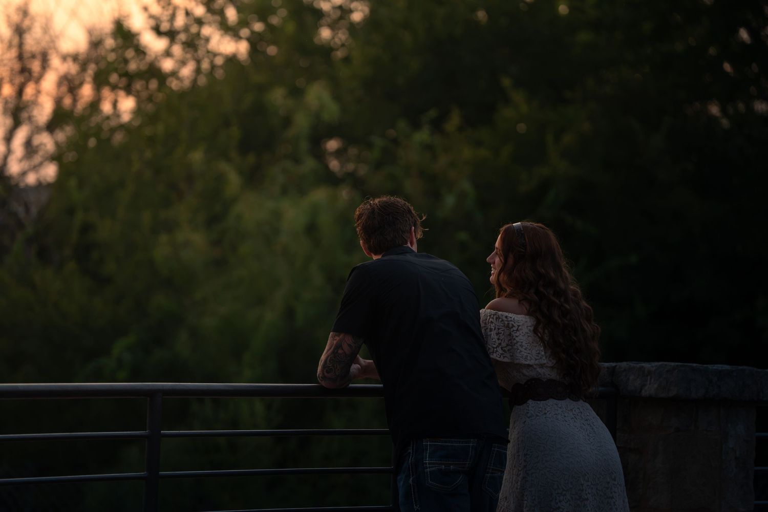 Couple standing together on a bridge at dusk, surrounded by lush green trees in soft evening light.