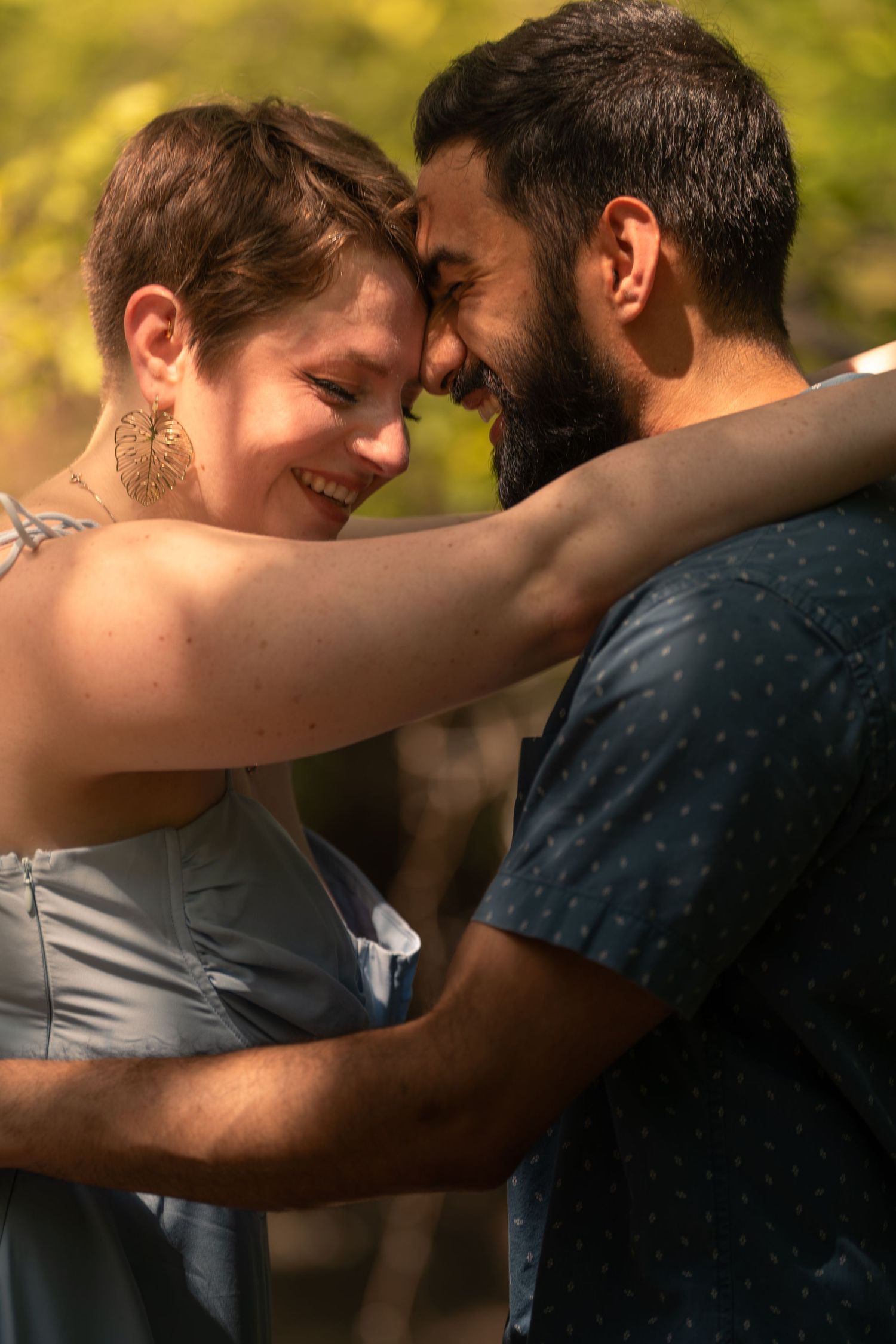 A smiling couple embraces tenderly outdoors, touching foreheads in a romantic moment surrounded by greenery.