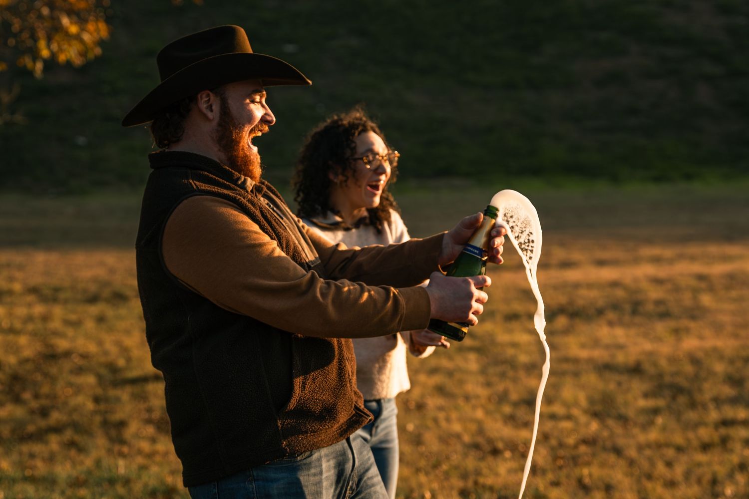 Couple laughing while popping champagne bottle outdoors in golden evening sunlight on grass.