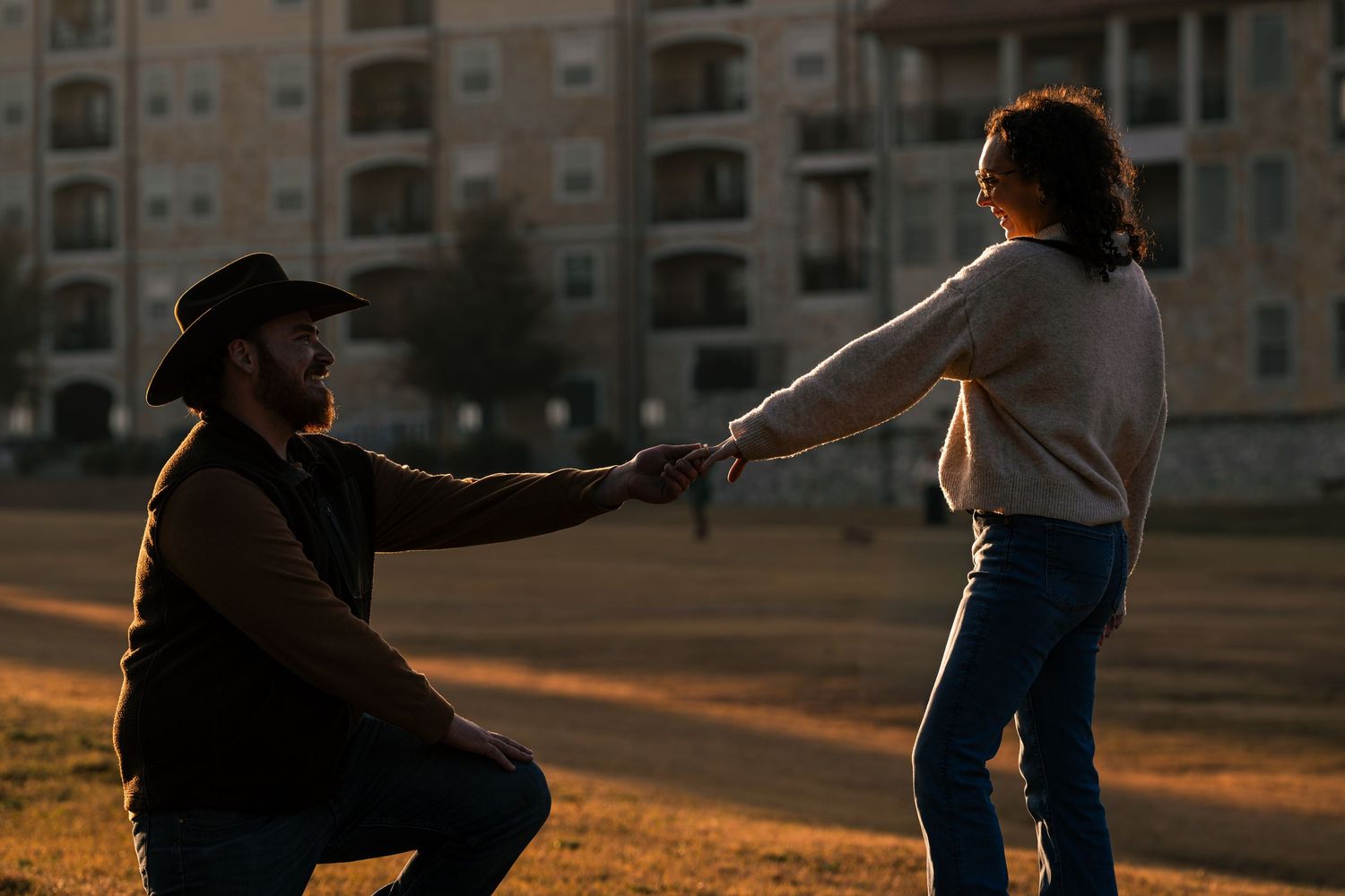Man in cowboy hat kneeling to propose to woman at romantic golden hour sunset.