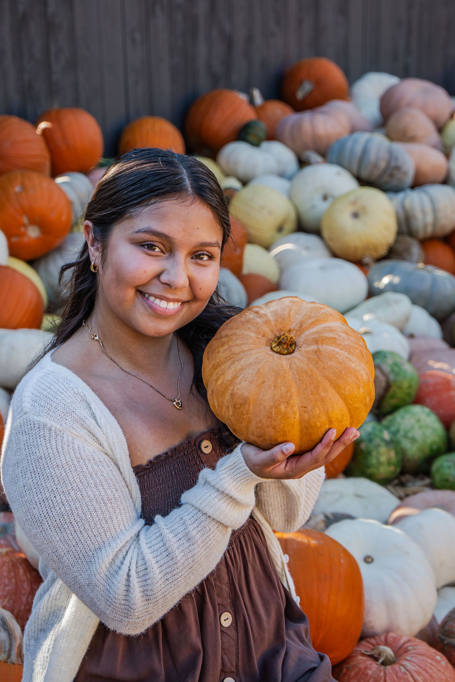 Smiling young woman holding an orange pumpkin in front of a colorful pumpkin display.