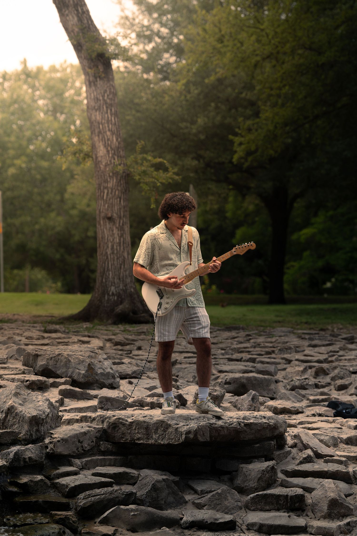 Young man playing electric guitar outdoors on rocky terrain with lush green trees in the background.