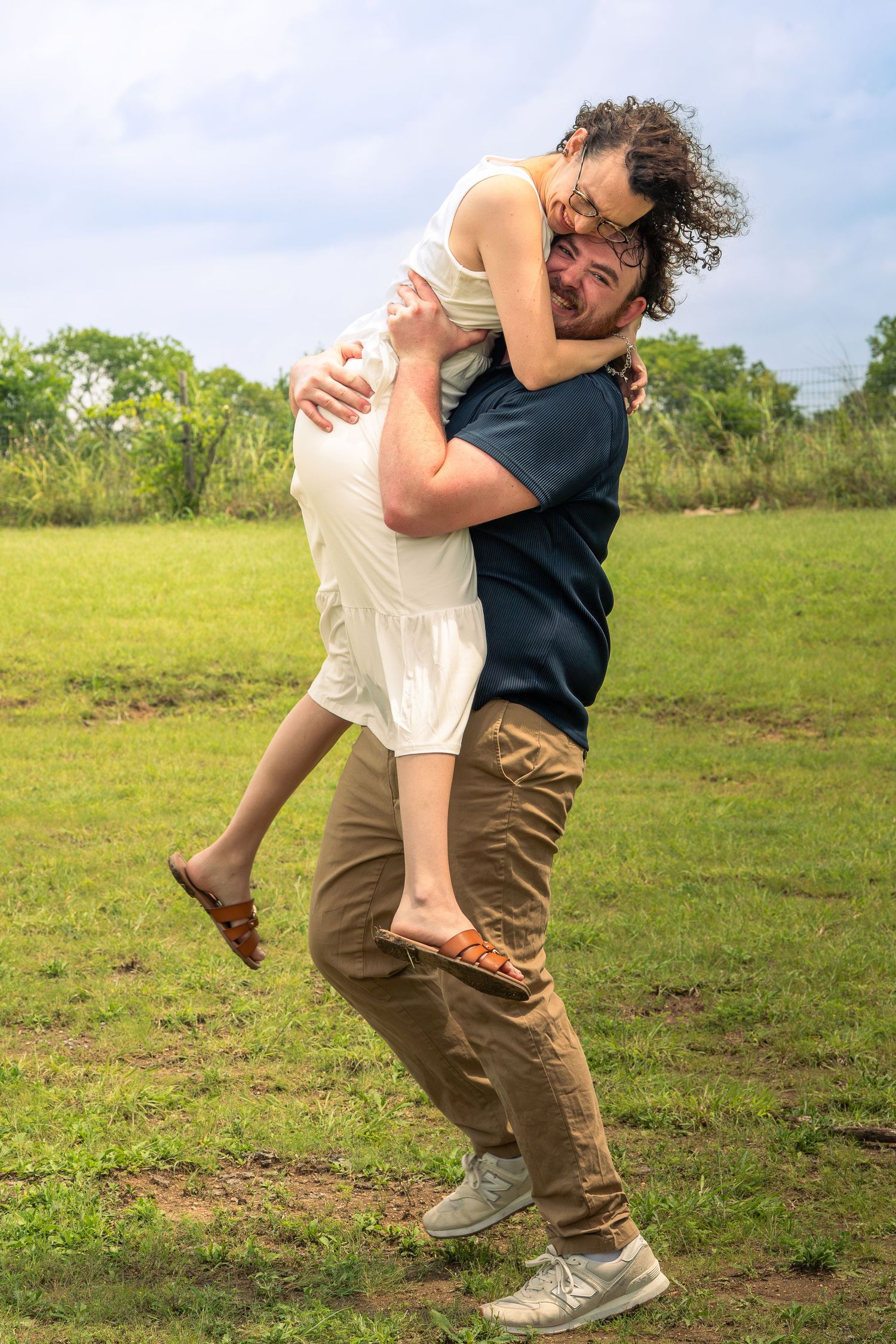 A man lifts and embraces a woman in a white dress outdoors on a green grassy field under a cloudy sky.