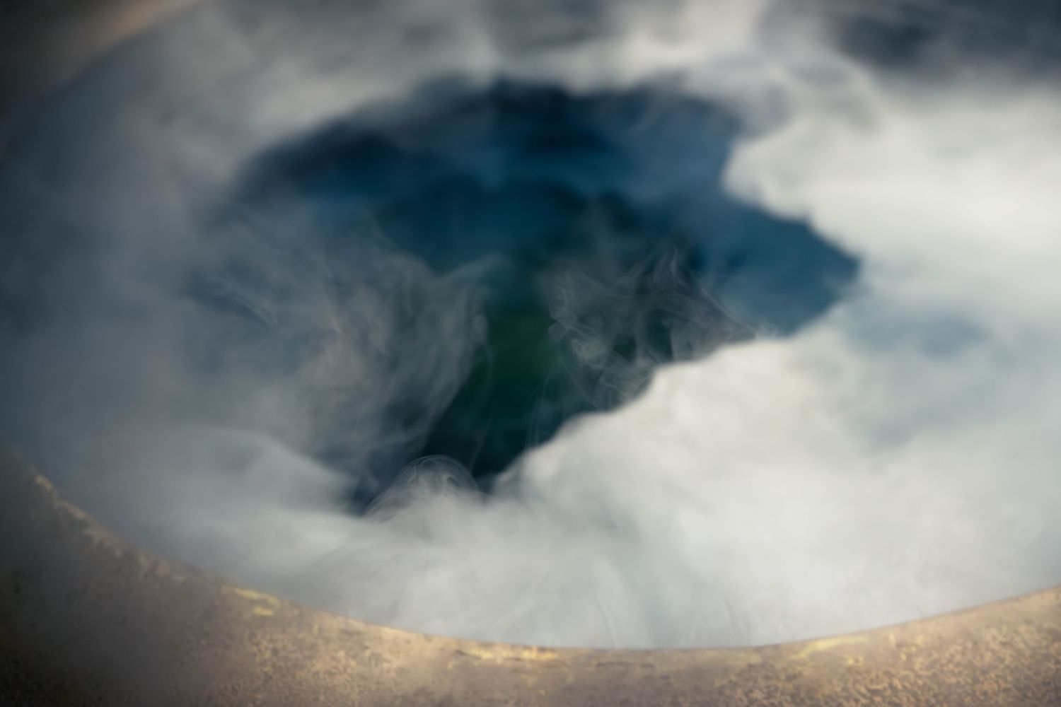 Aerial view of a steaming geothermal hot spring pool with misty clouds and deep blue-green water.