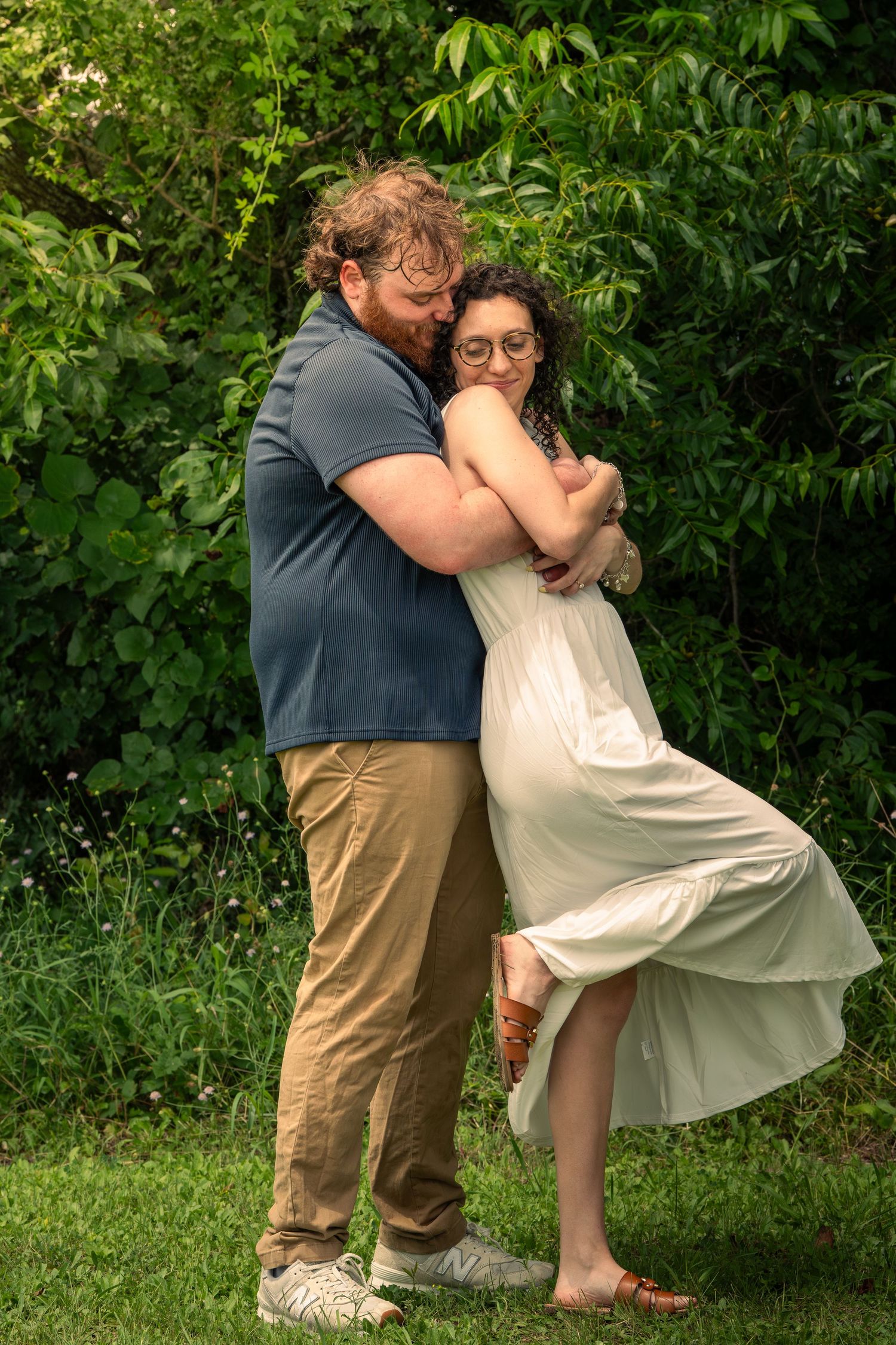 A couple embraces tenderly in a lush green garden, woman in white dress, man in blue shirt and tan pants.