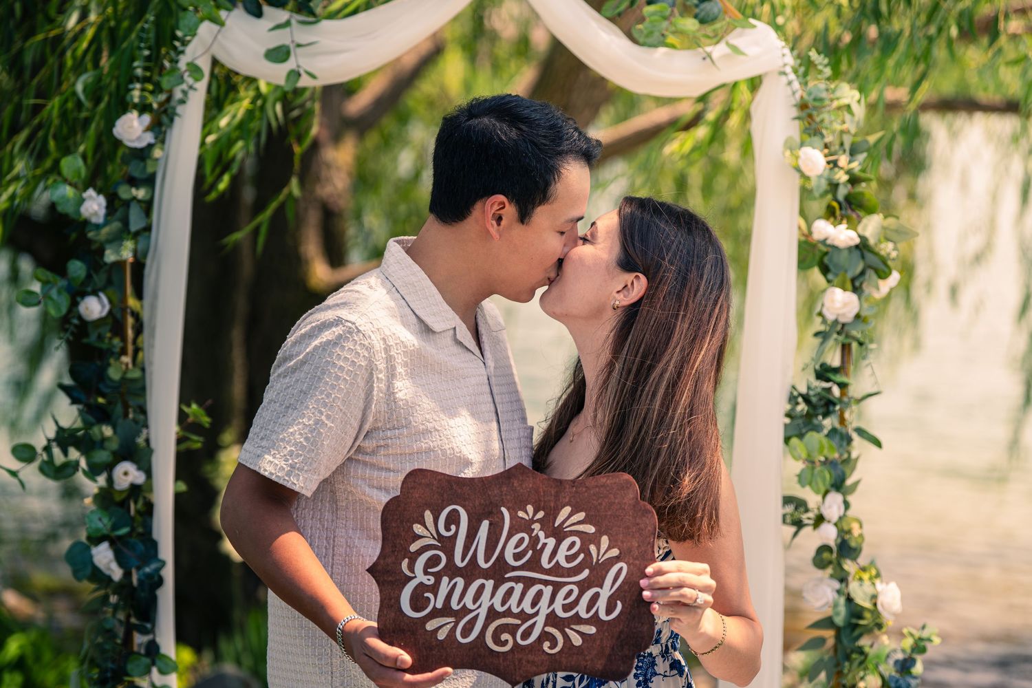 Couple kissing under floral arch holding We're Engaged sign at outdoor engagement photo shoot.