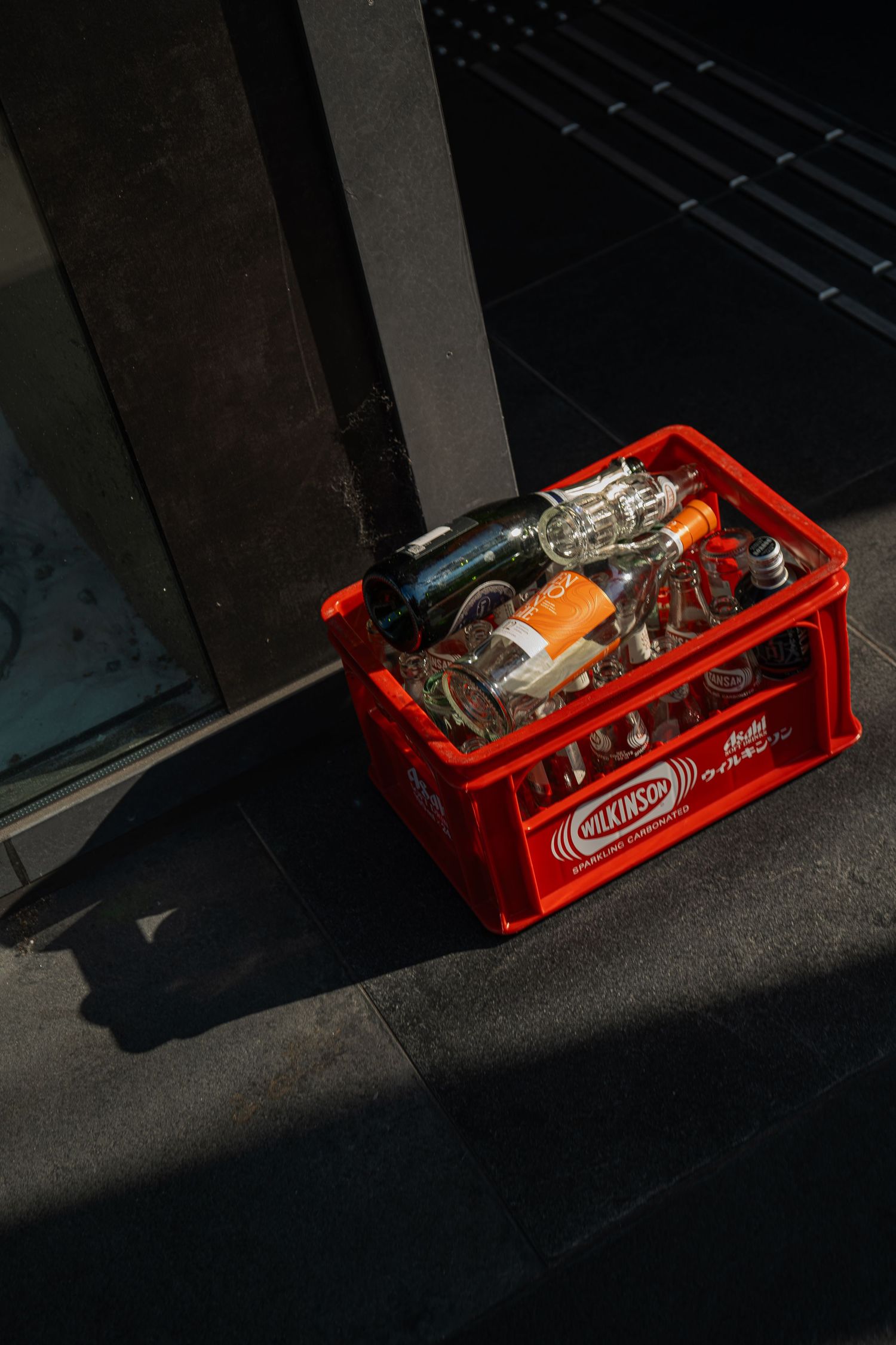 Red plastic Milkivop bottle crate filled with assorted empty glass bottles on sunlit pavement.