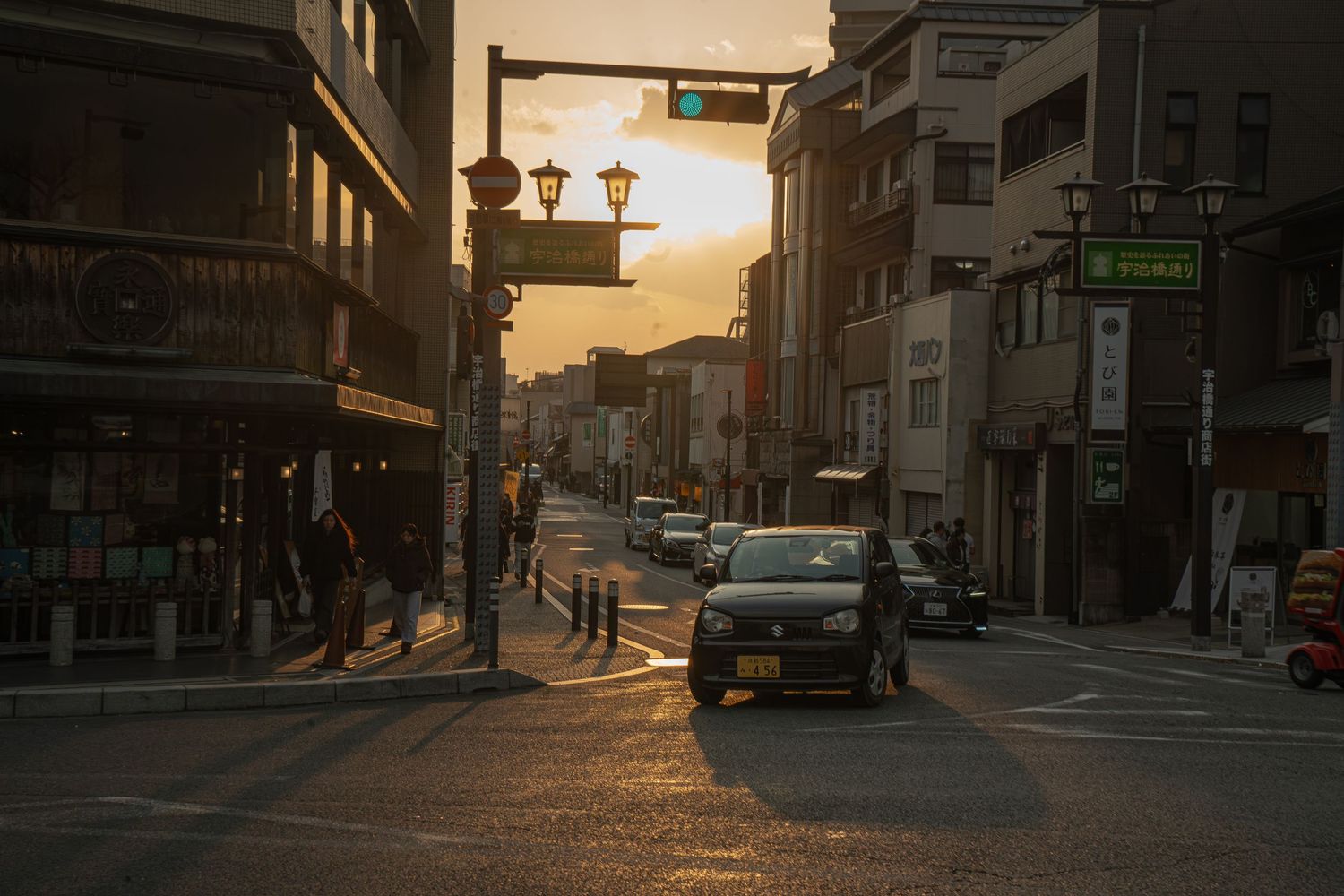 Golden sunset over a quiet Japanese city street with cars and pedestrians at an intersection.