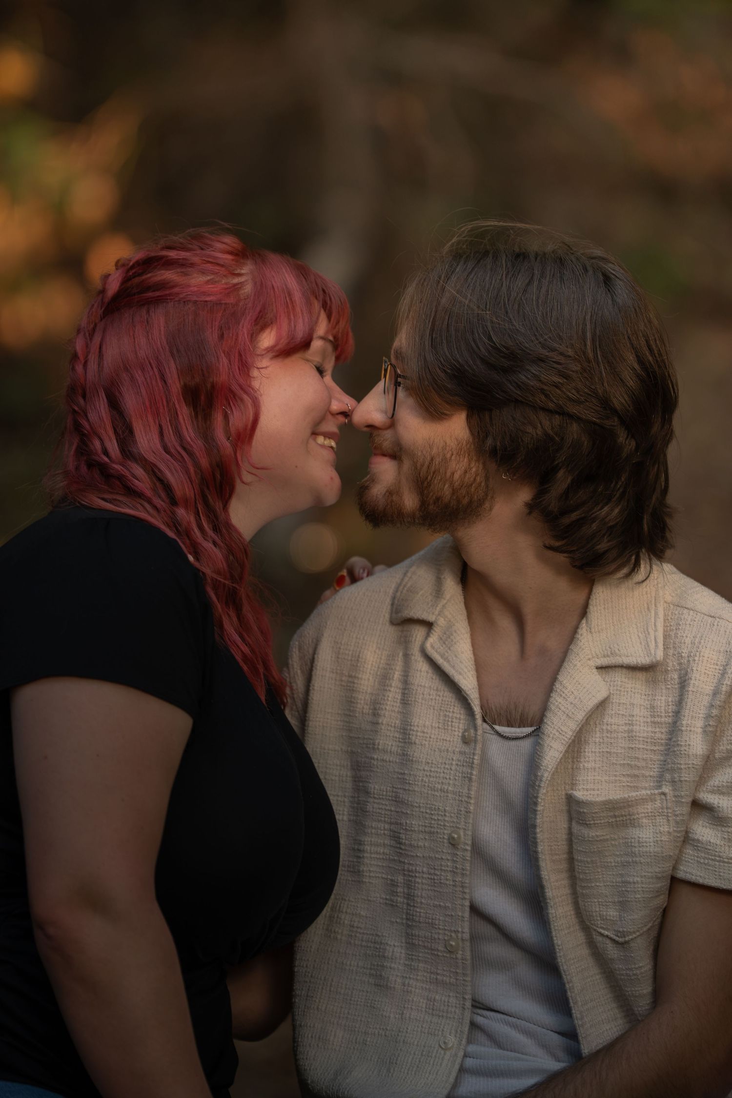 A couple with red-haired woman and bearded man in glasses share a tender nose-to-nose moment outdoors in warm autumn light.
