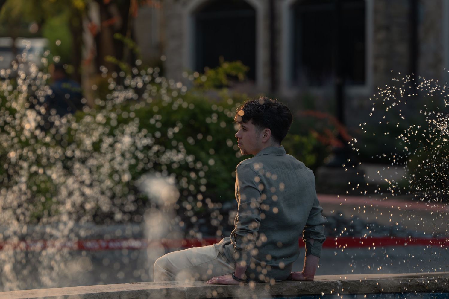 Young man sitting by a fountain outdoors in warm golden light, water splashing around him.